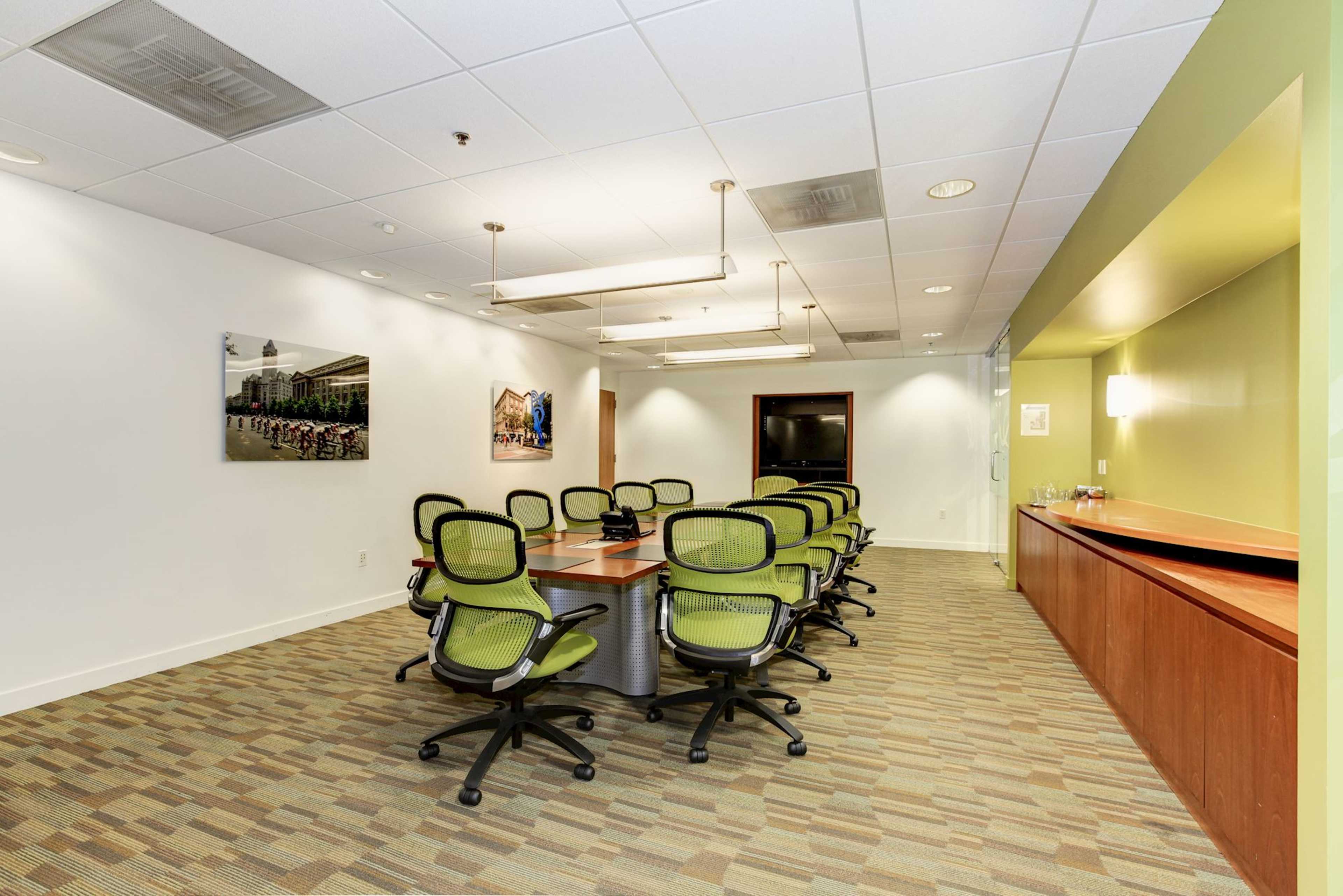A modern conference room with a large table surrounded by ten green office chairs, adjacent to a wooden counter and a wall featuring a photograph.