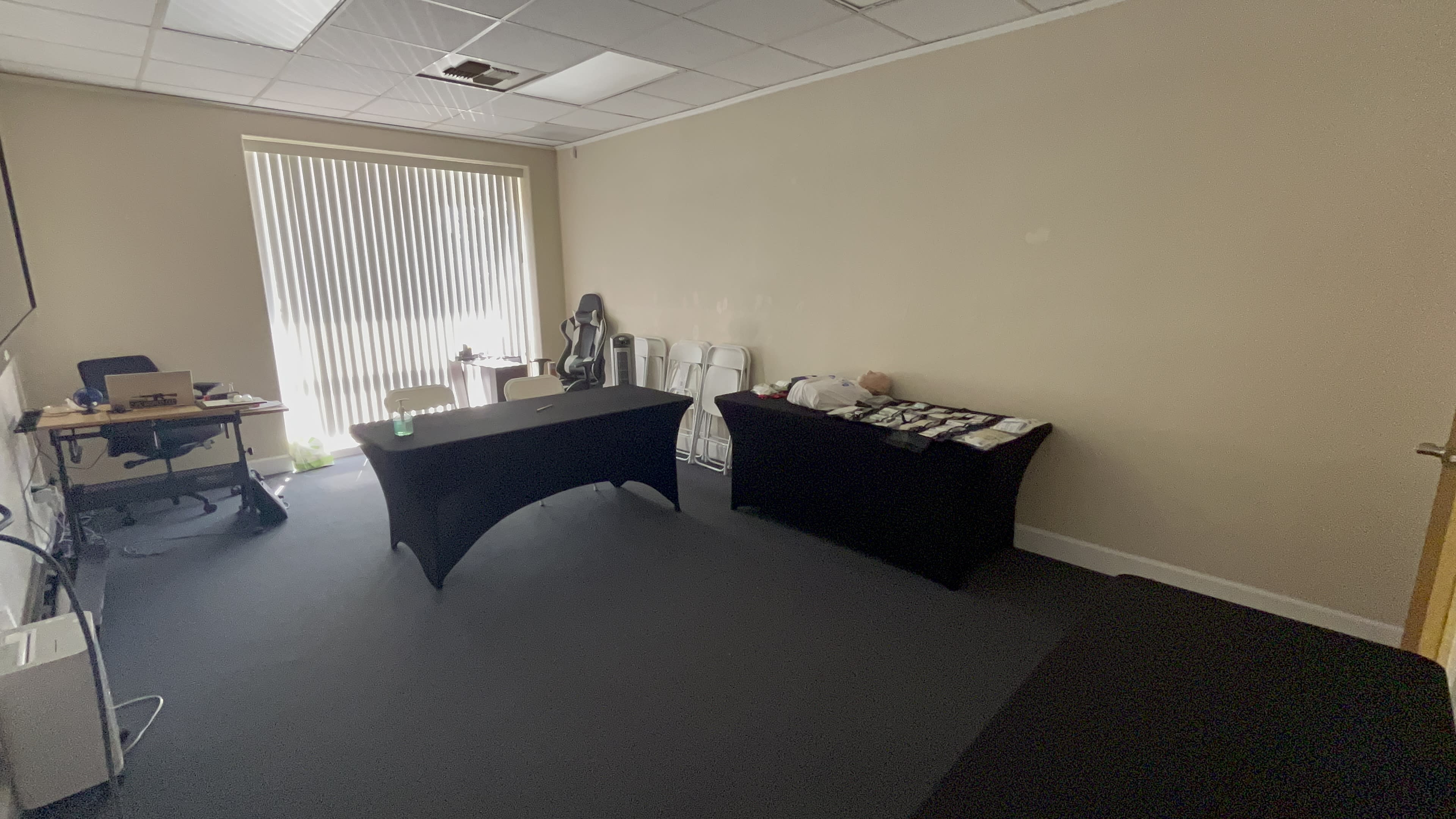 A sparsely furnished room with two tables covered in black cloth and a row of white chairs against one wall, illuminated by natural light from a large window.