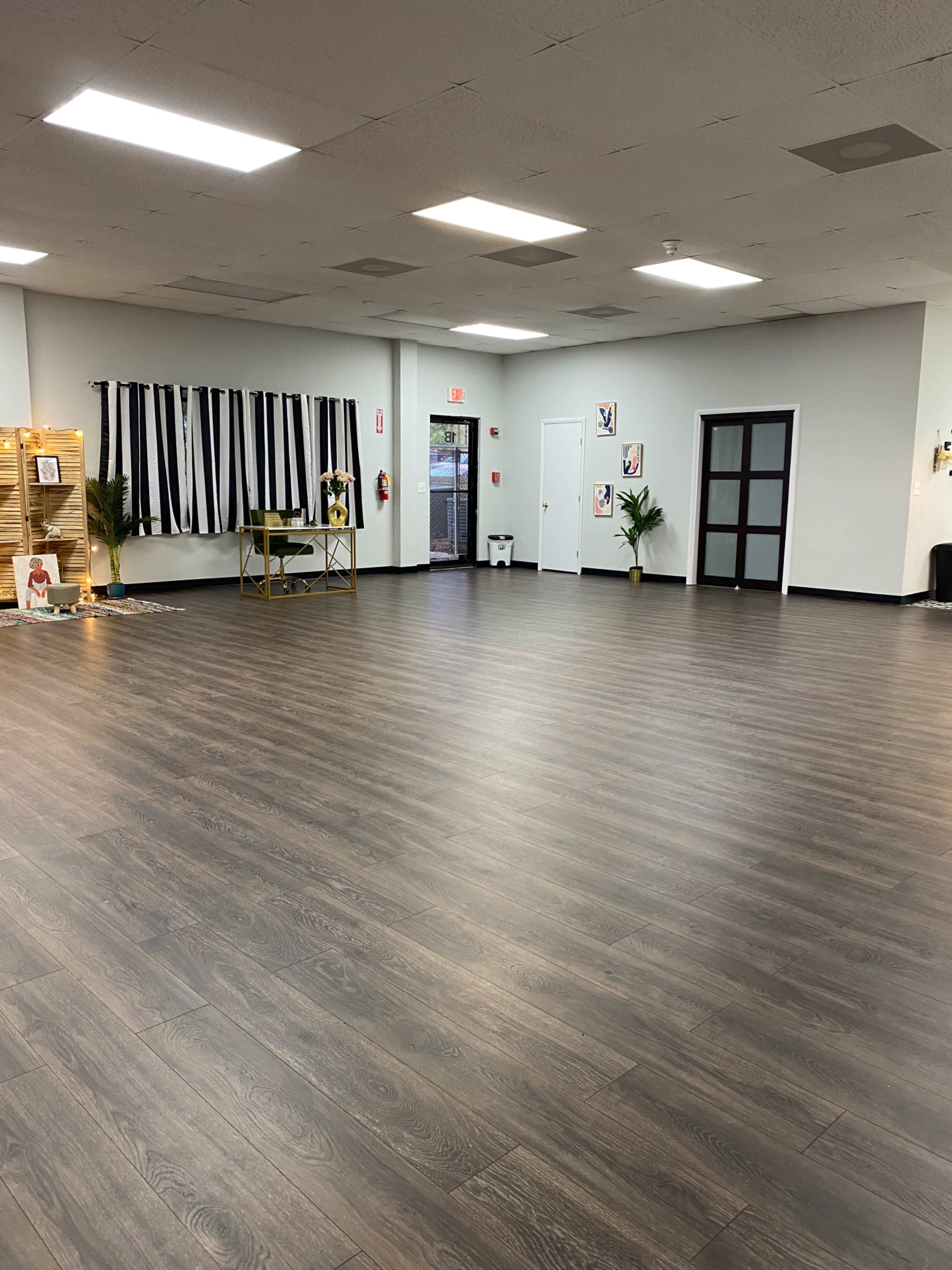 The image shows a spacious interior room with wood flooring, large windows, and a minimalistic design featuring chairs and decorative plants.