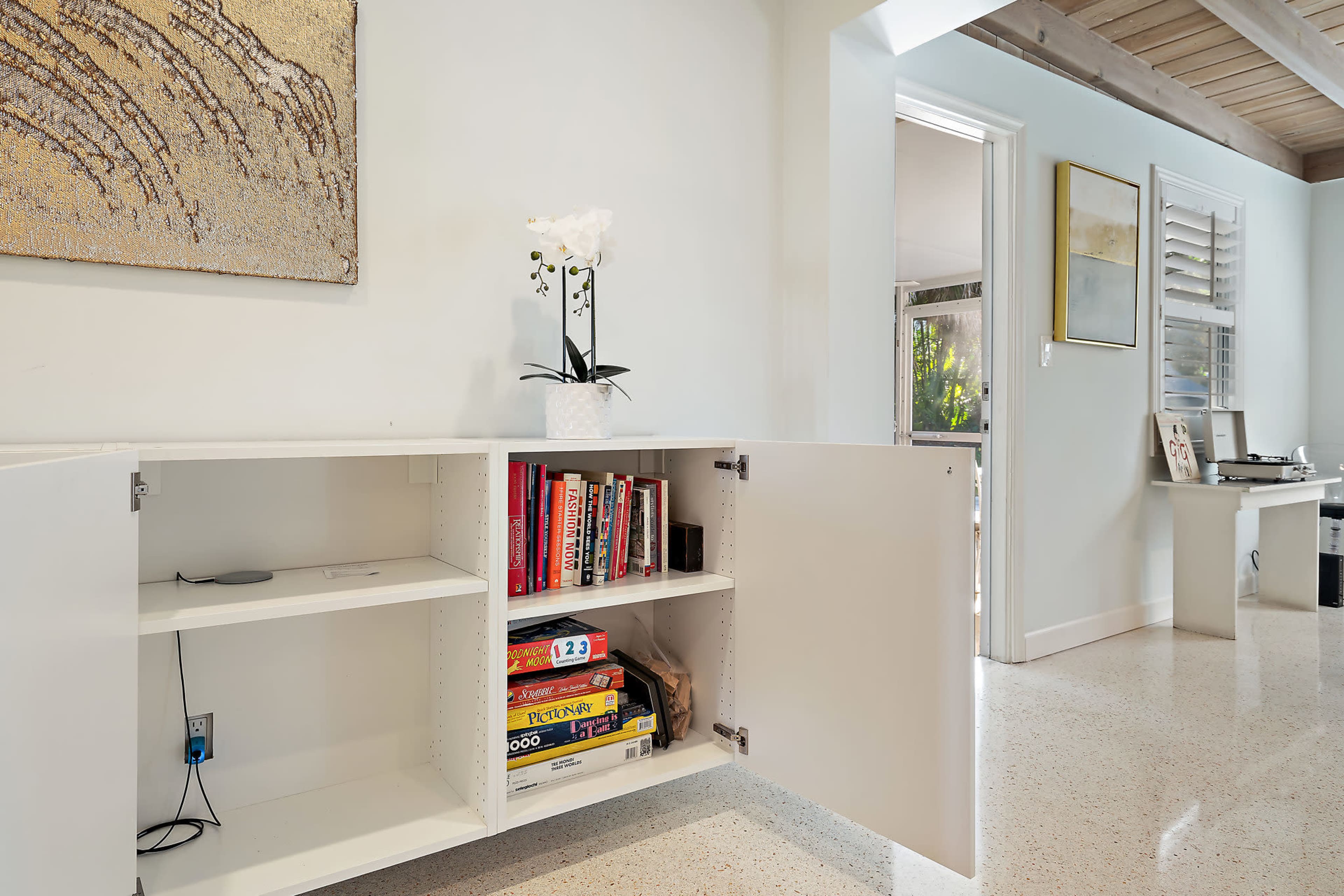 A modern living space features a white cabinet with open doors displaying books and board games, beside a plant and artwork on the wall.