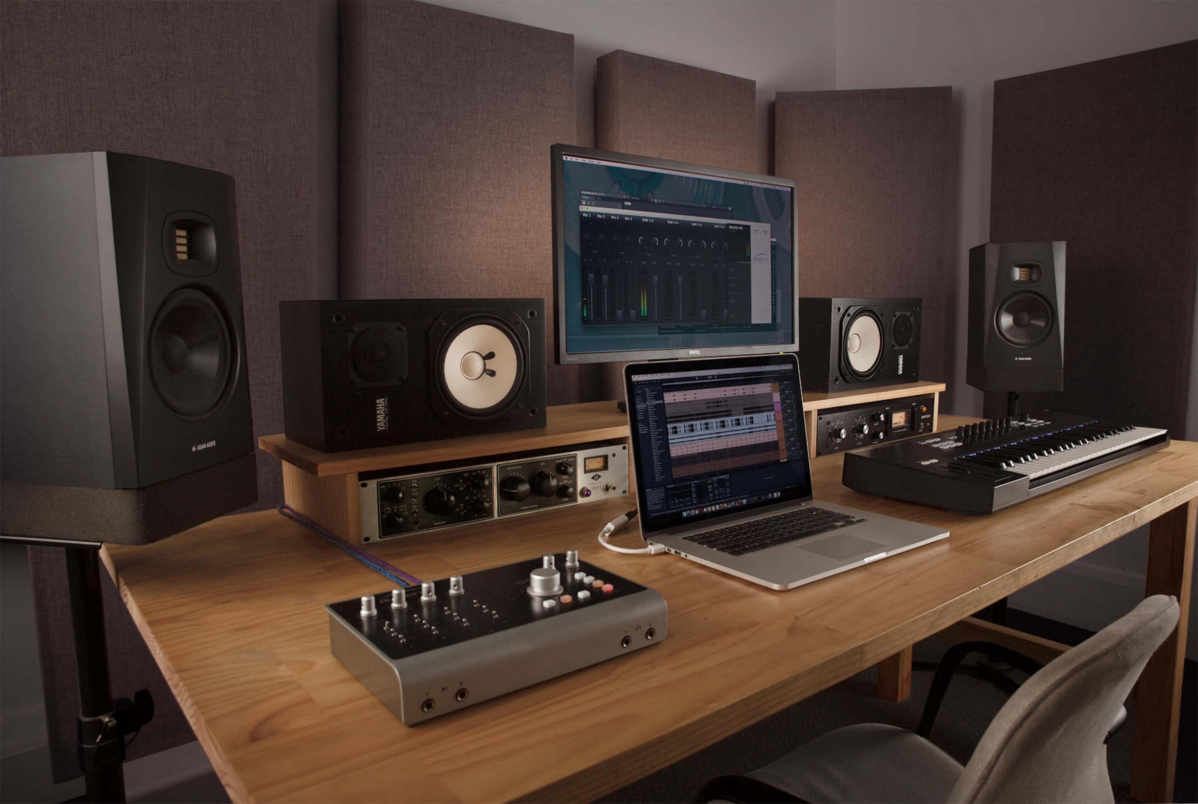A wooden desk in a music studio features a laptop, audio interface, MIDI keyboard, and two studio monitors, with acoustic panels on the wall behind.