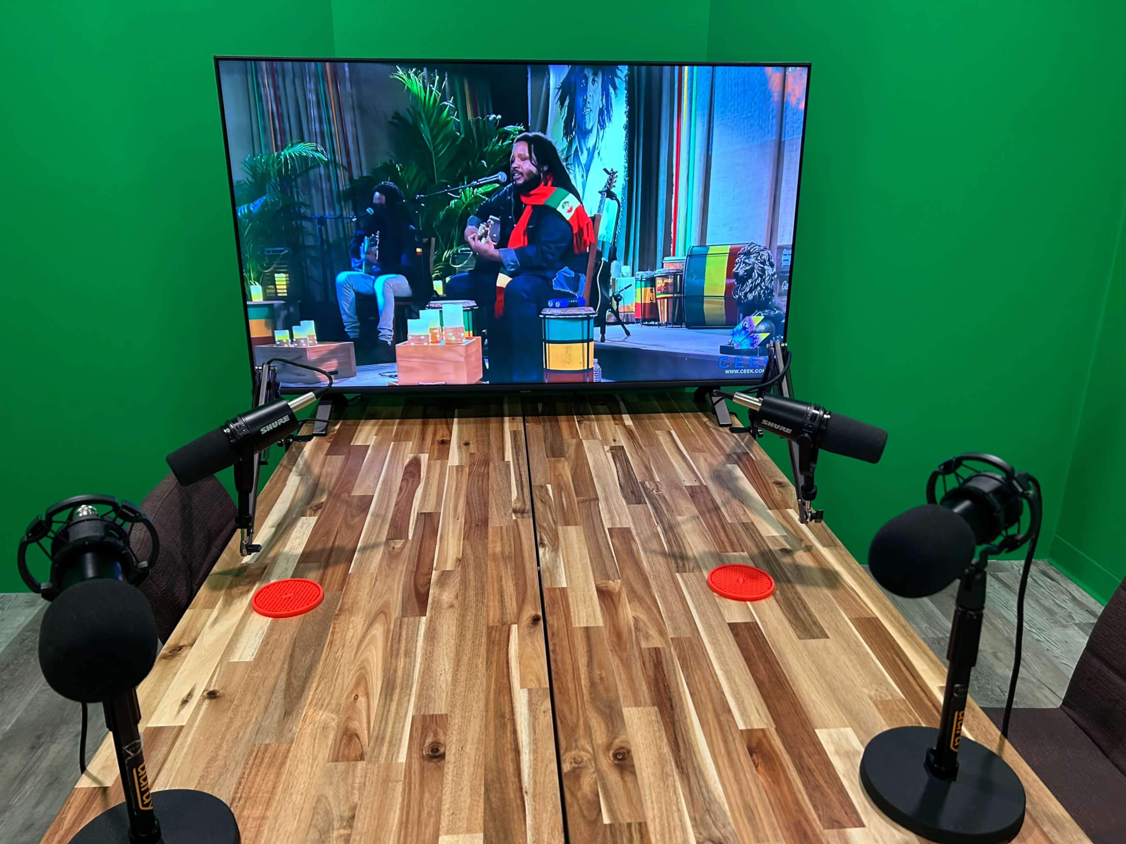 A wooden table with microphones is set up in front of a large TV screen displaying a performance scene.