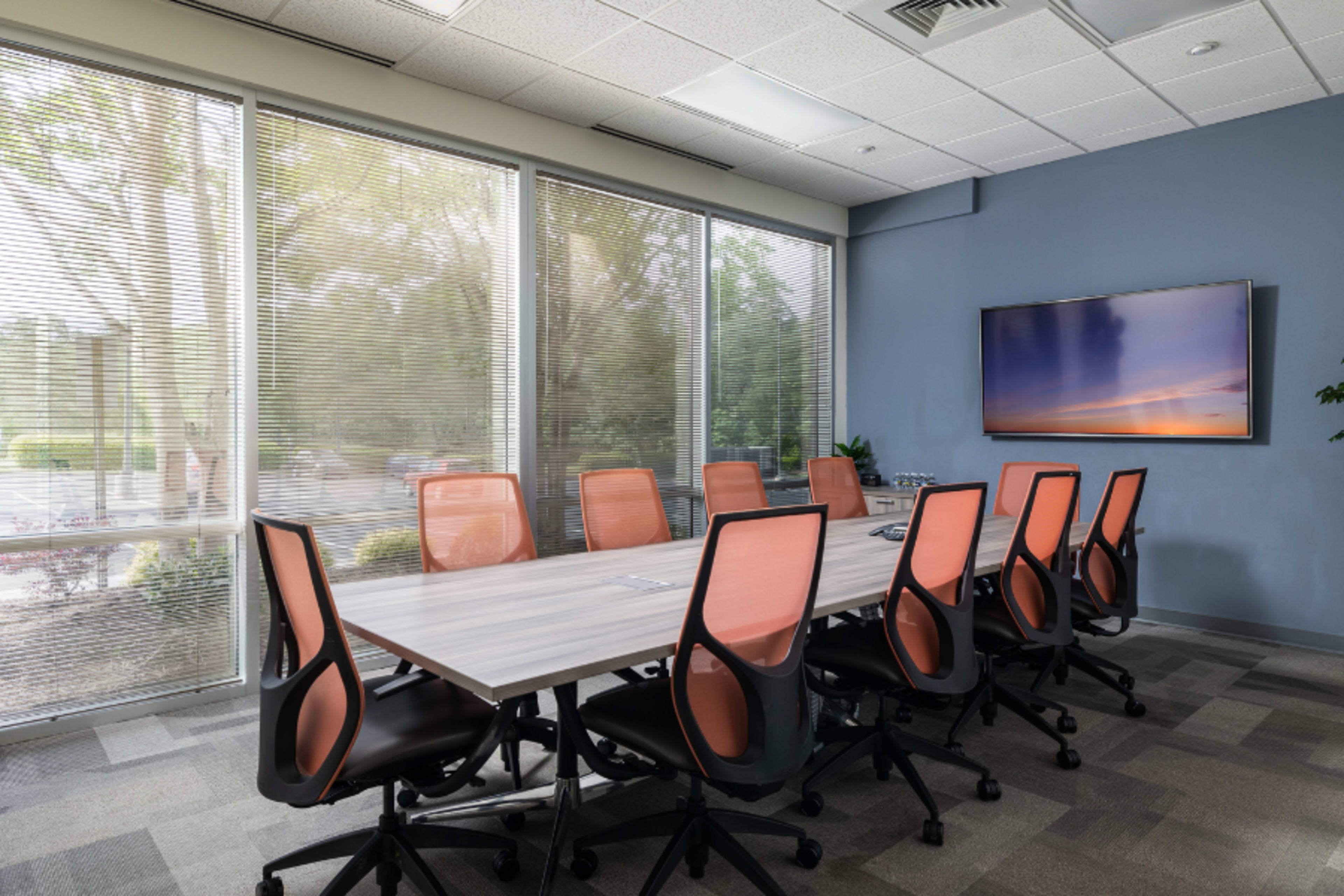 A modern conference room features a long table surrounded by eight ergonomic chairs, with large windows allowing natural light and a television mounted on a blue wall.
