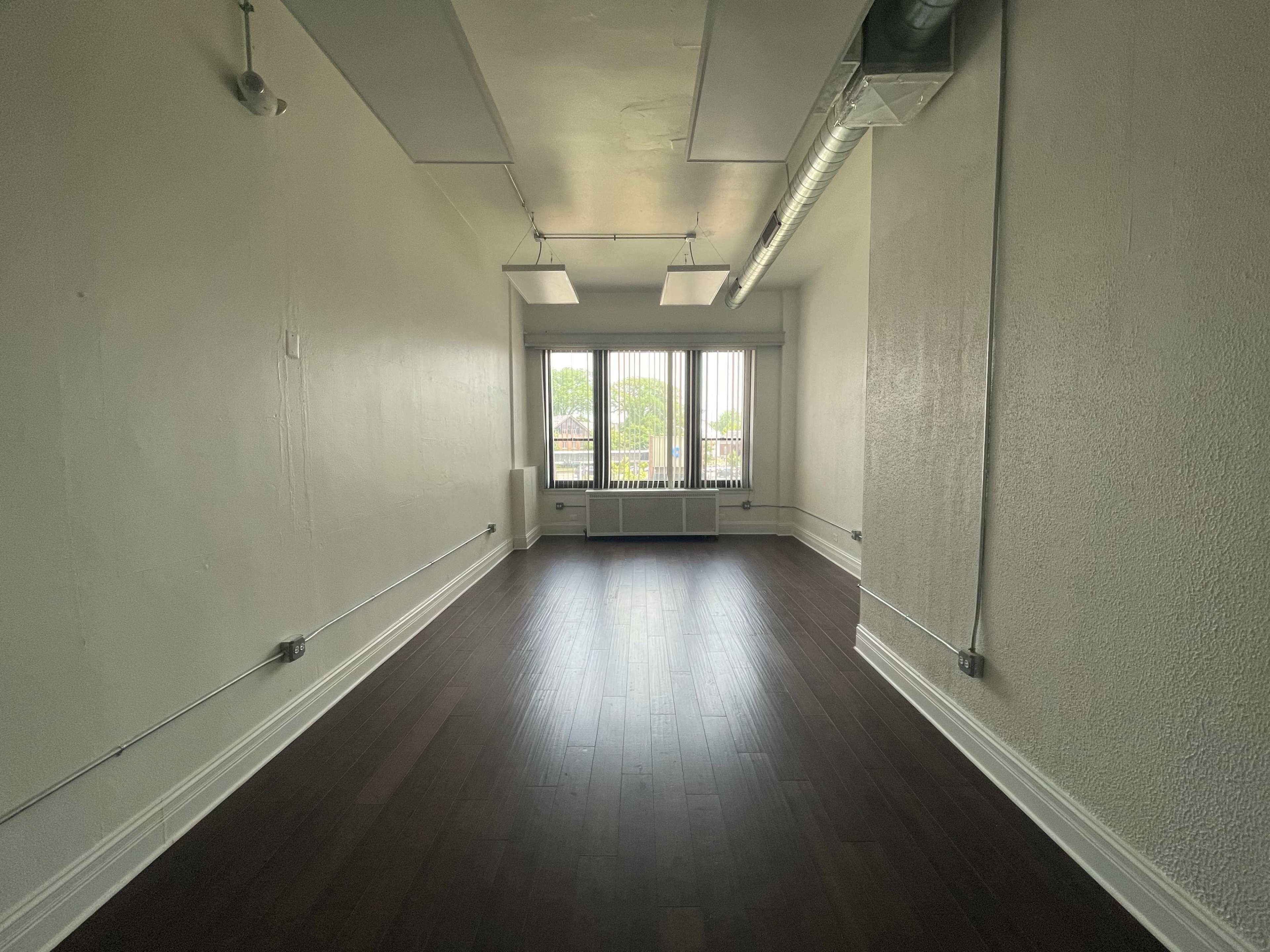An empty room with hardwood floors features large windows facing a view of trees outside.
