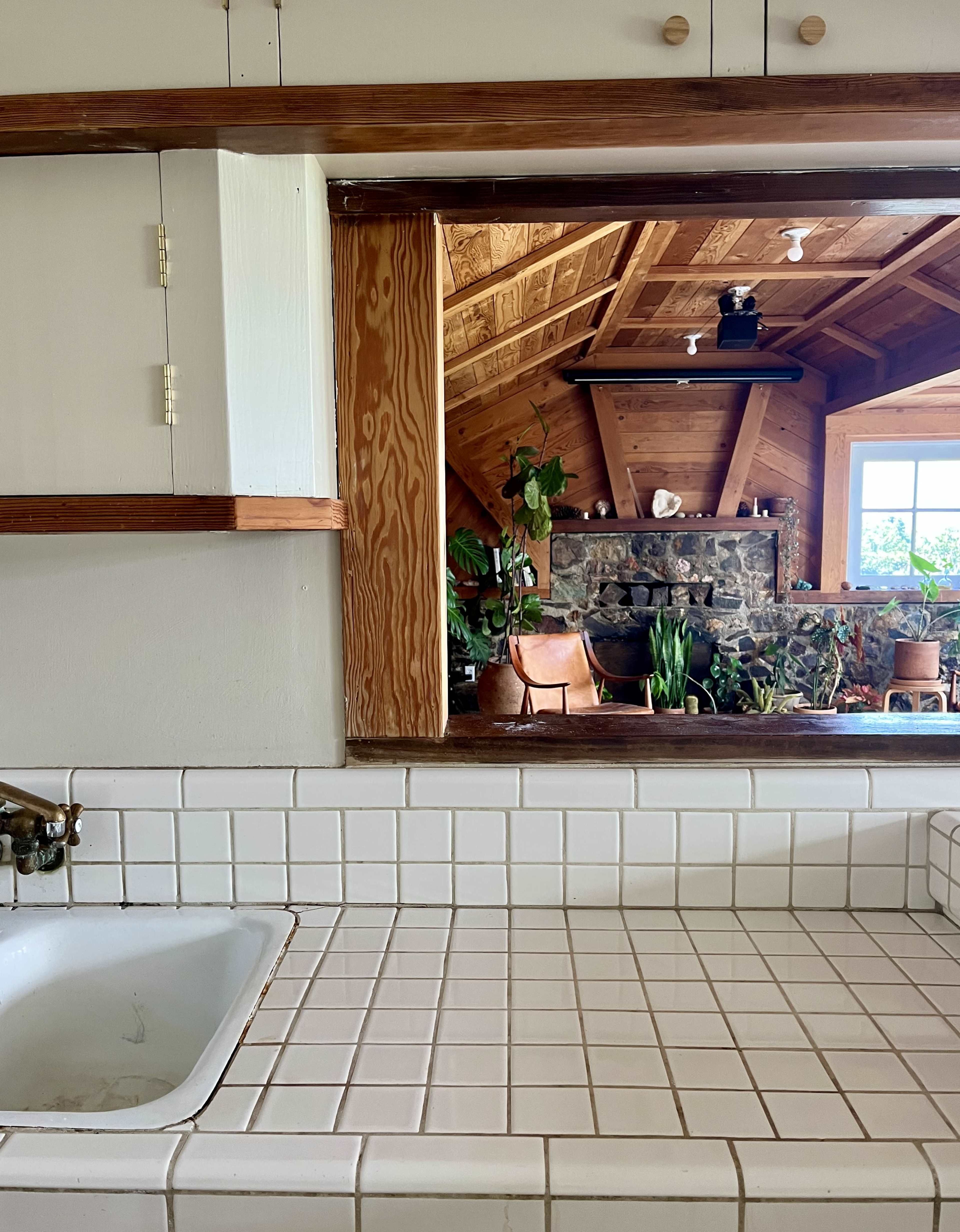 A kitchen sink area with tiled countertops and an open window leading to a space featuring a stone fireplace and numerous plants.