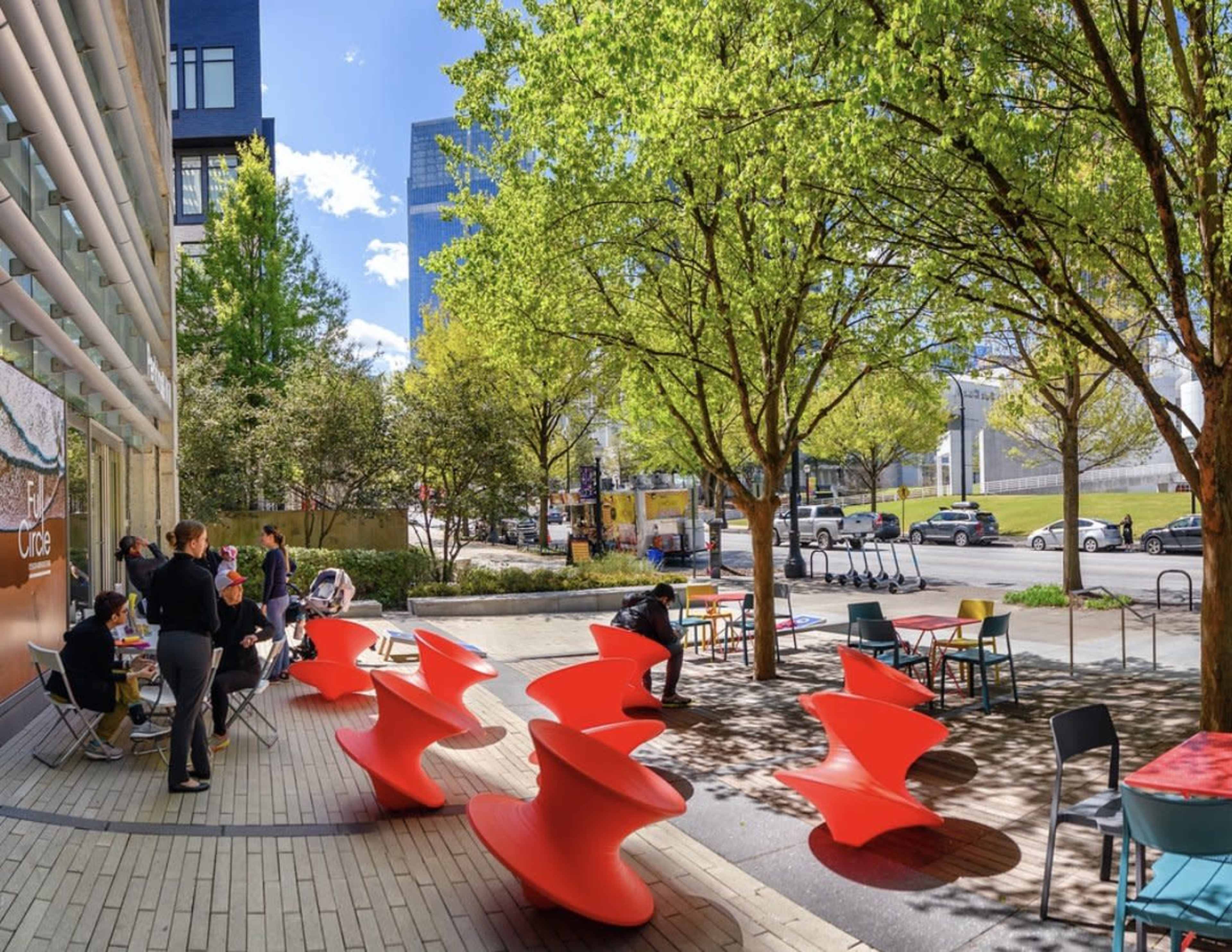 A modern outdoor space with colorful red seating, trees, and people enjoying the area near a building and street.