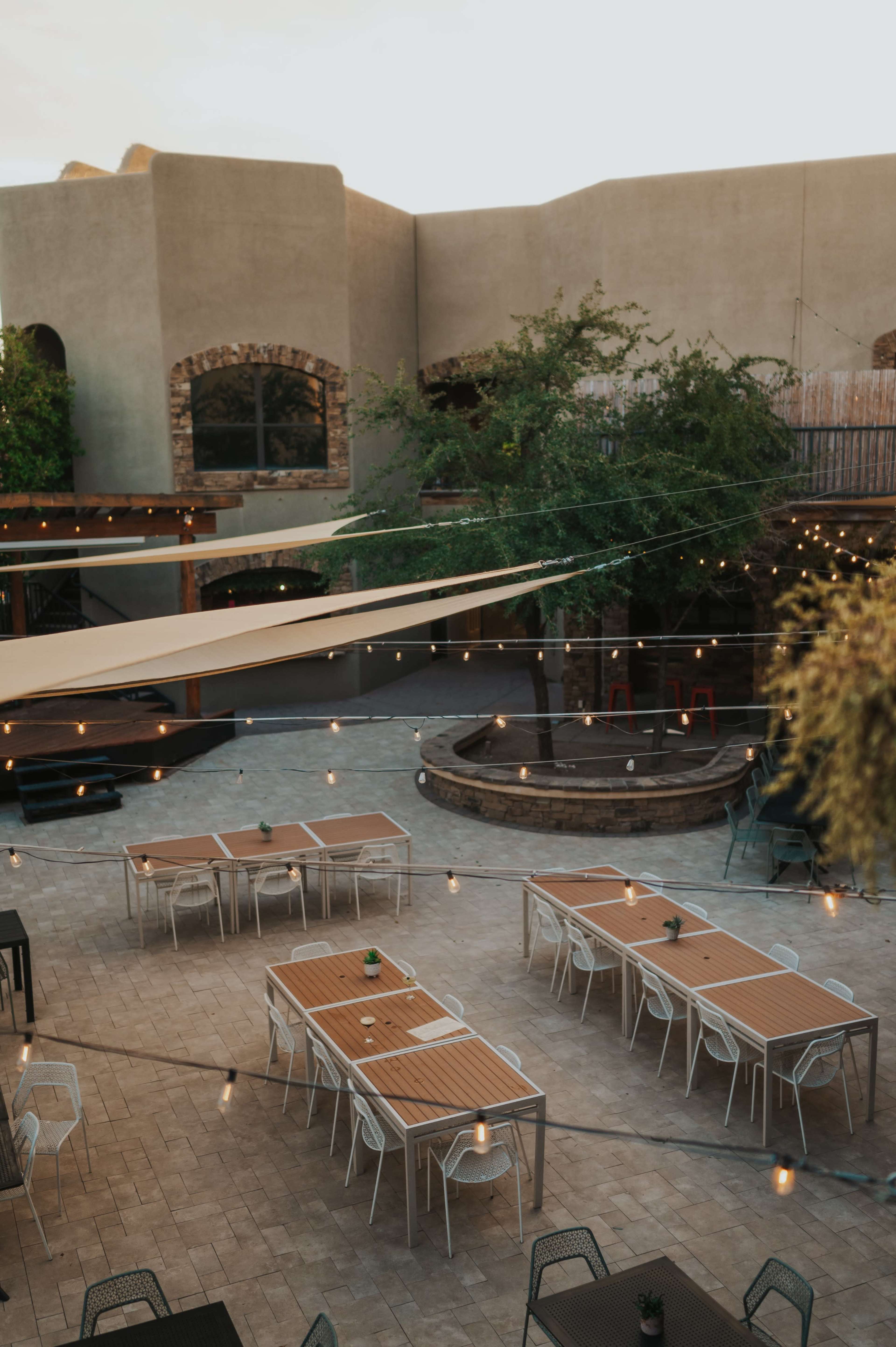 The image shows an outdoor dining area with multiple tables arranged under string lights, surrounded by walls and greenery.