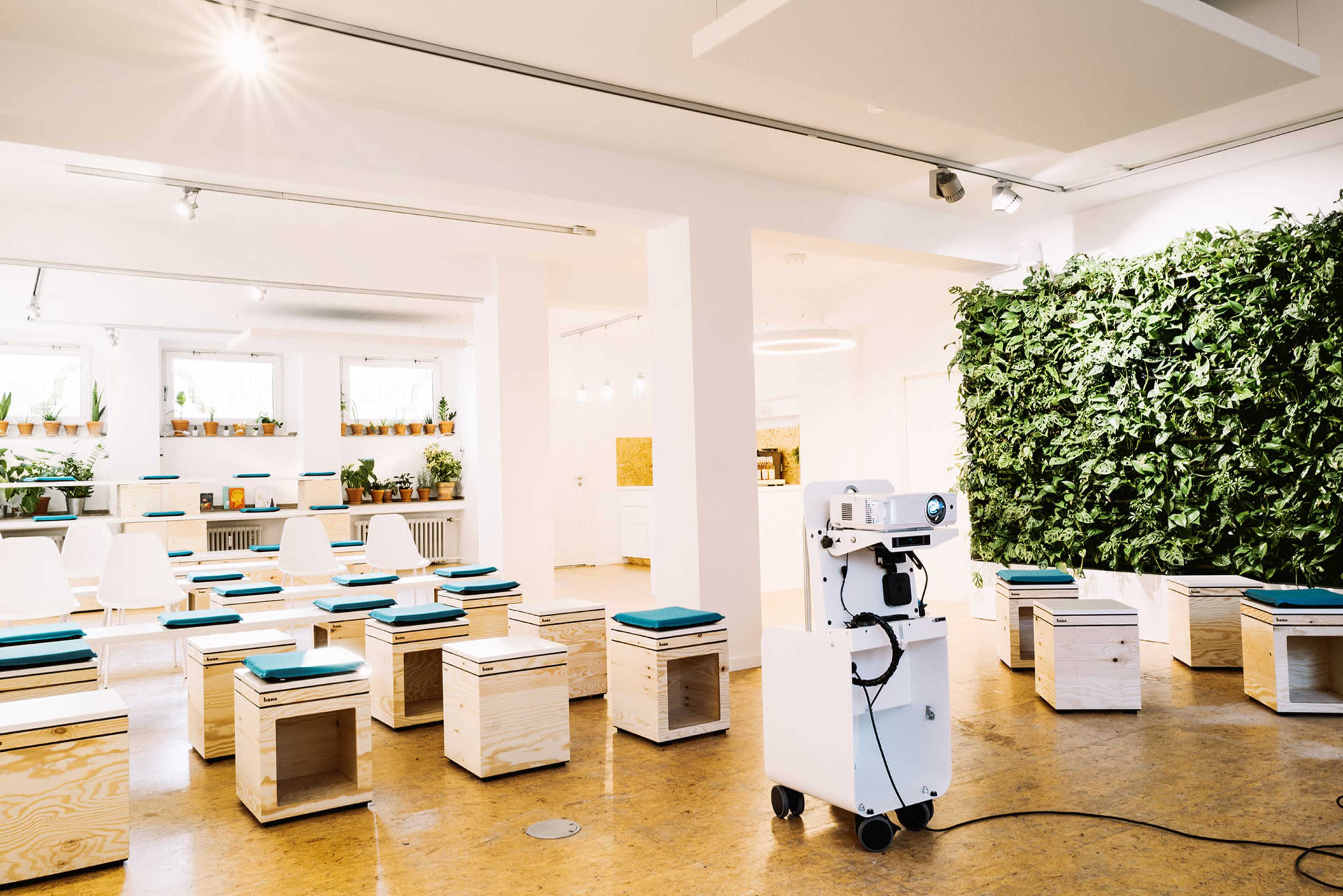 A bright, minimalist presentation room with wooden stools, a projector on a stand, and a wall covered in greenery.