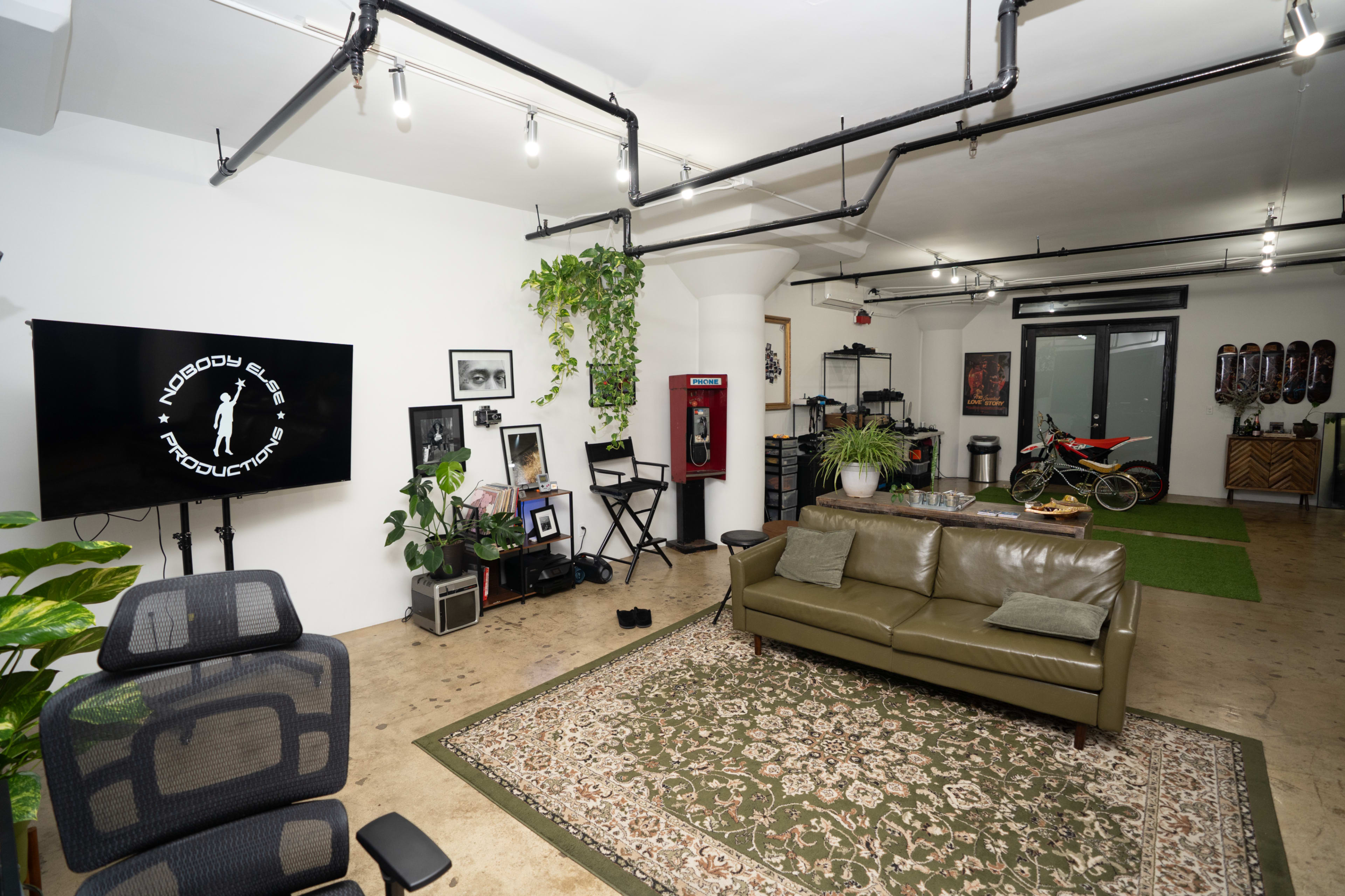The image shows a spacious urban studio with a green couch, various plants, a television mounted on the wall, and a motorcycle displayed in the background.