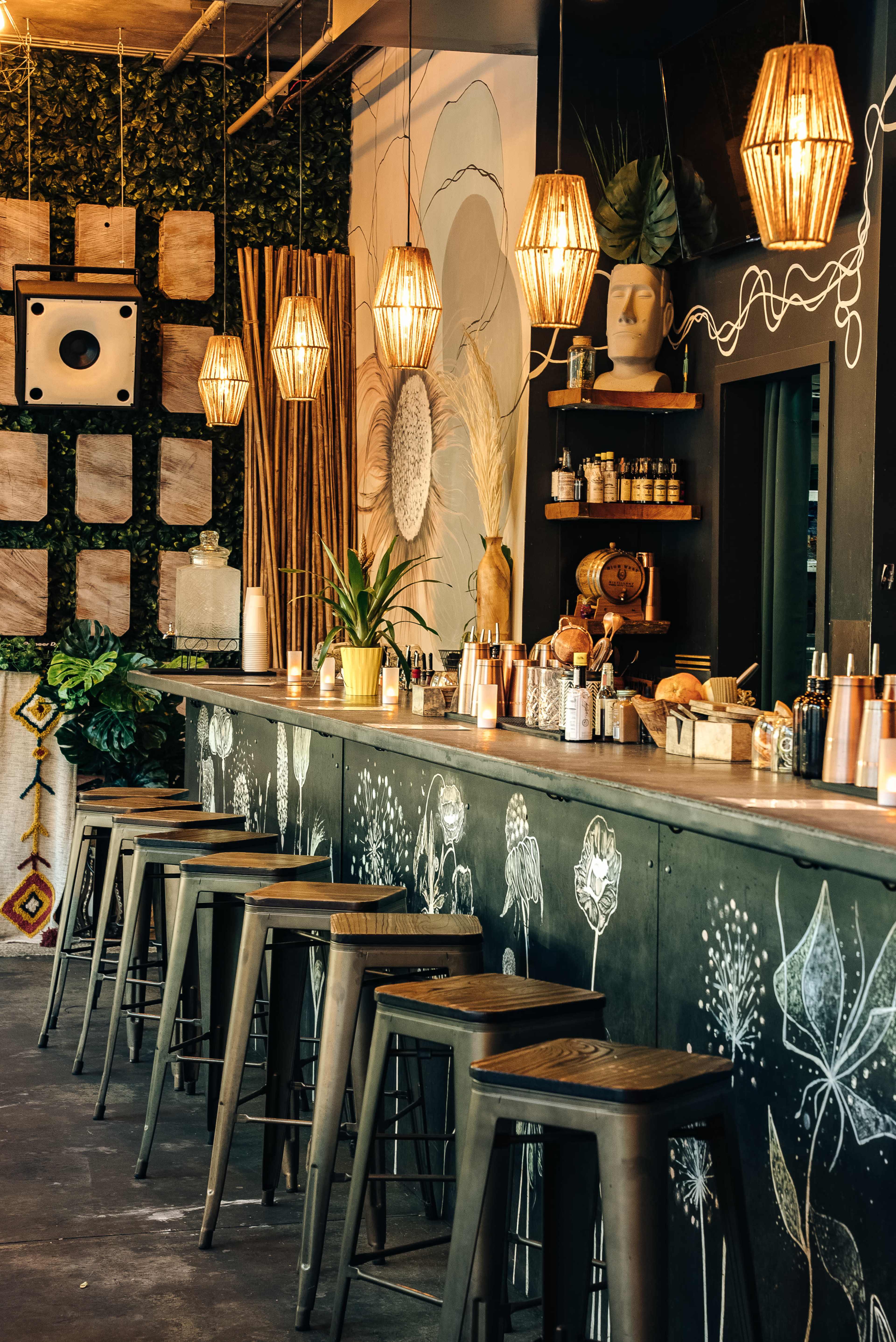 The image shows a stylish bar area with a dark countertop, metal stools, decorative lighting, and floral wall art.