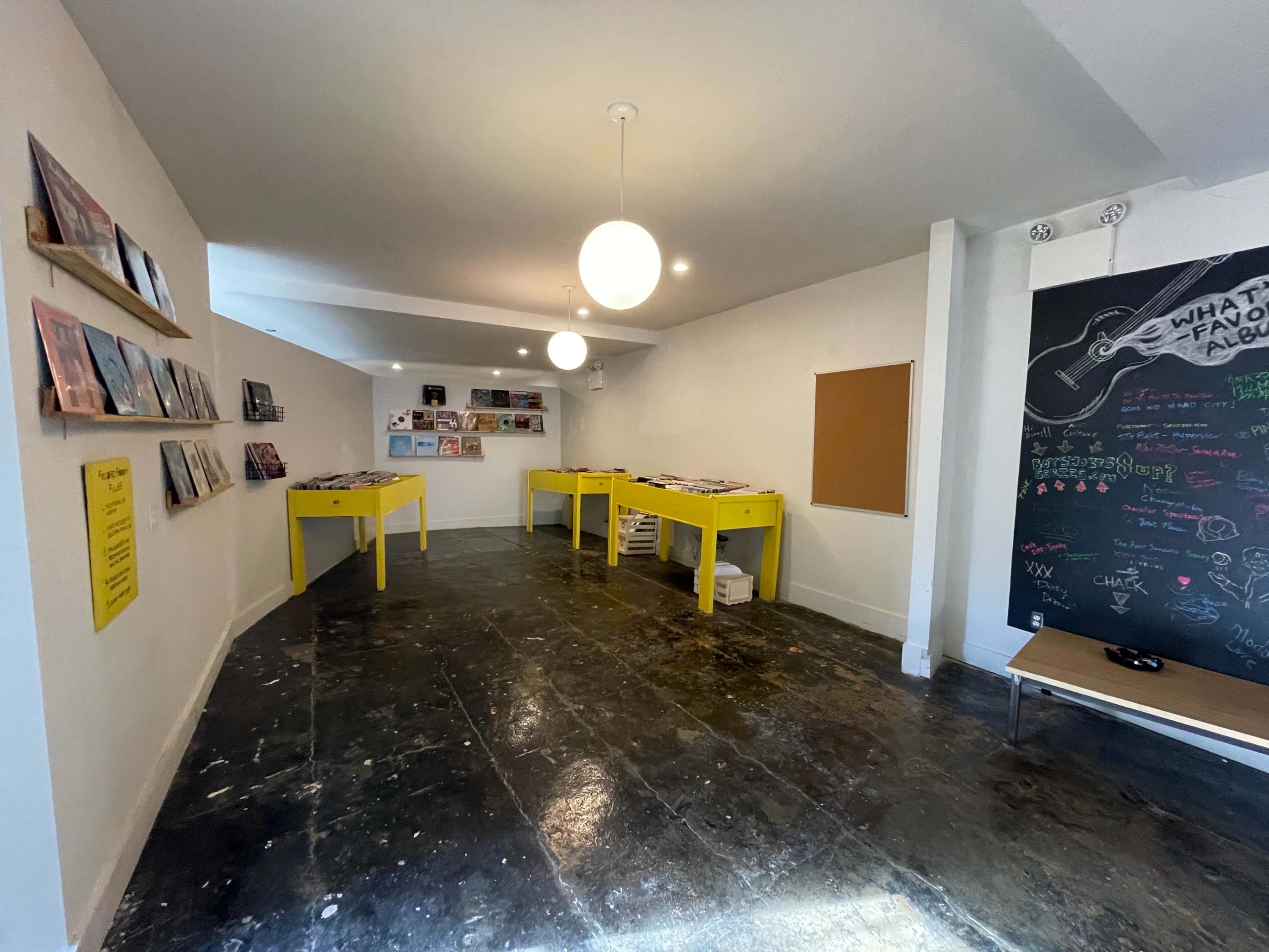 A minimalistic room with yellow tables displaying records, a wall featuring vinyl album covers, and a chalkboard with colorful drawings and writing.
