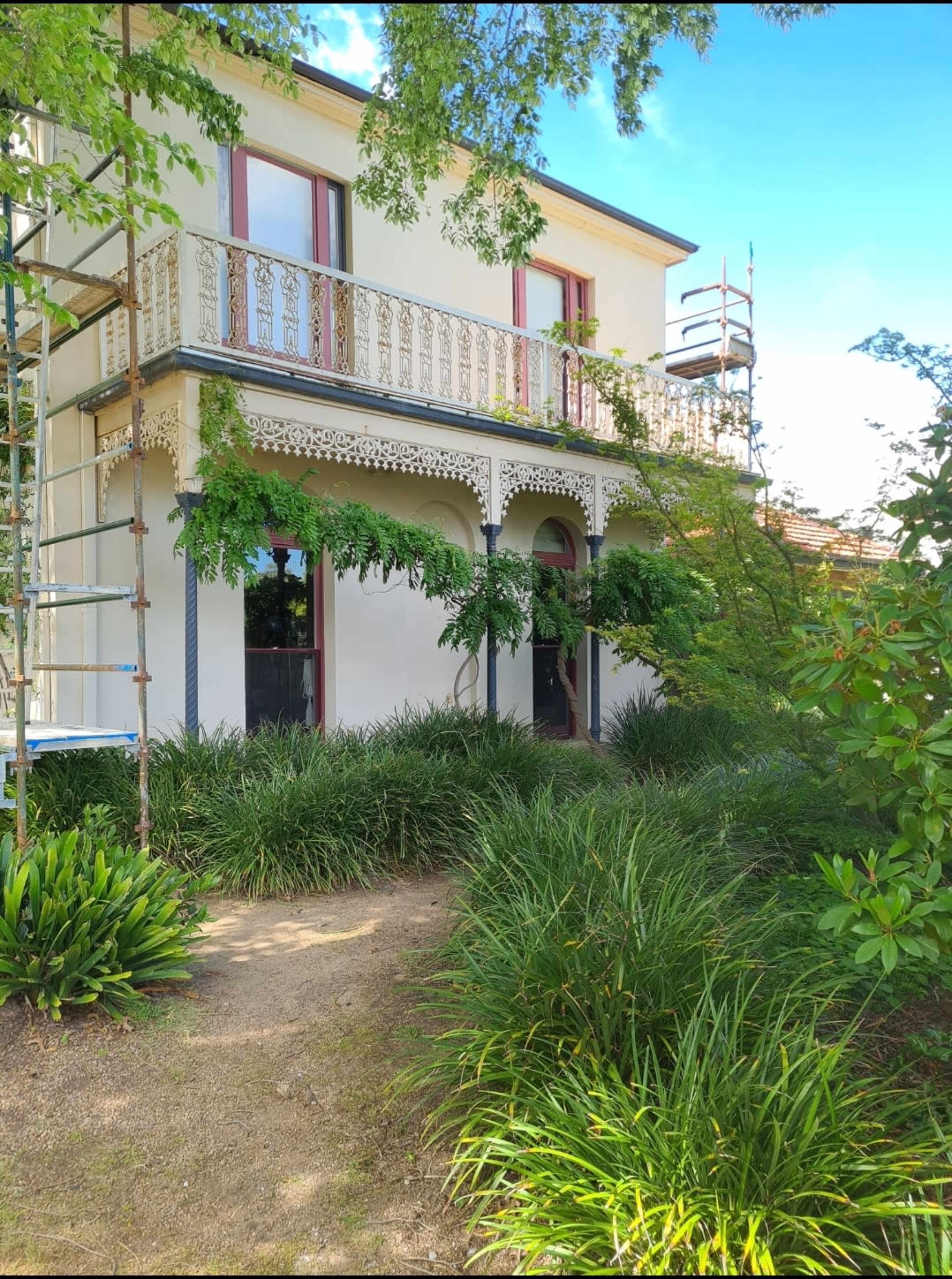 A two-story house with a balcony surrounded by lush greenery and scaffolding on one side.