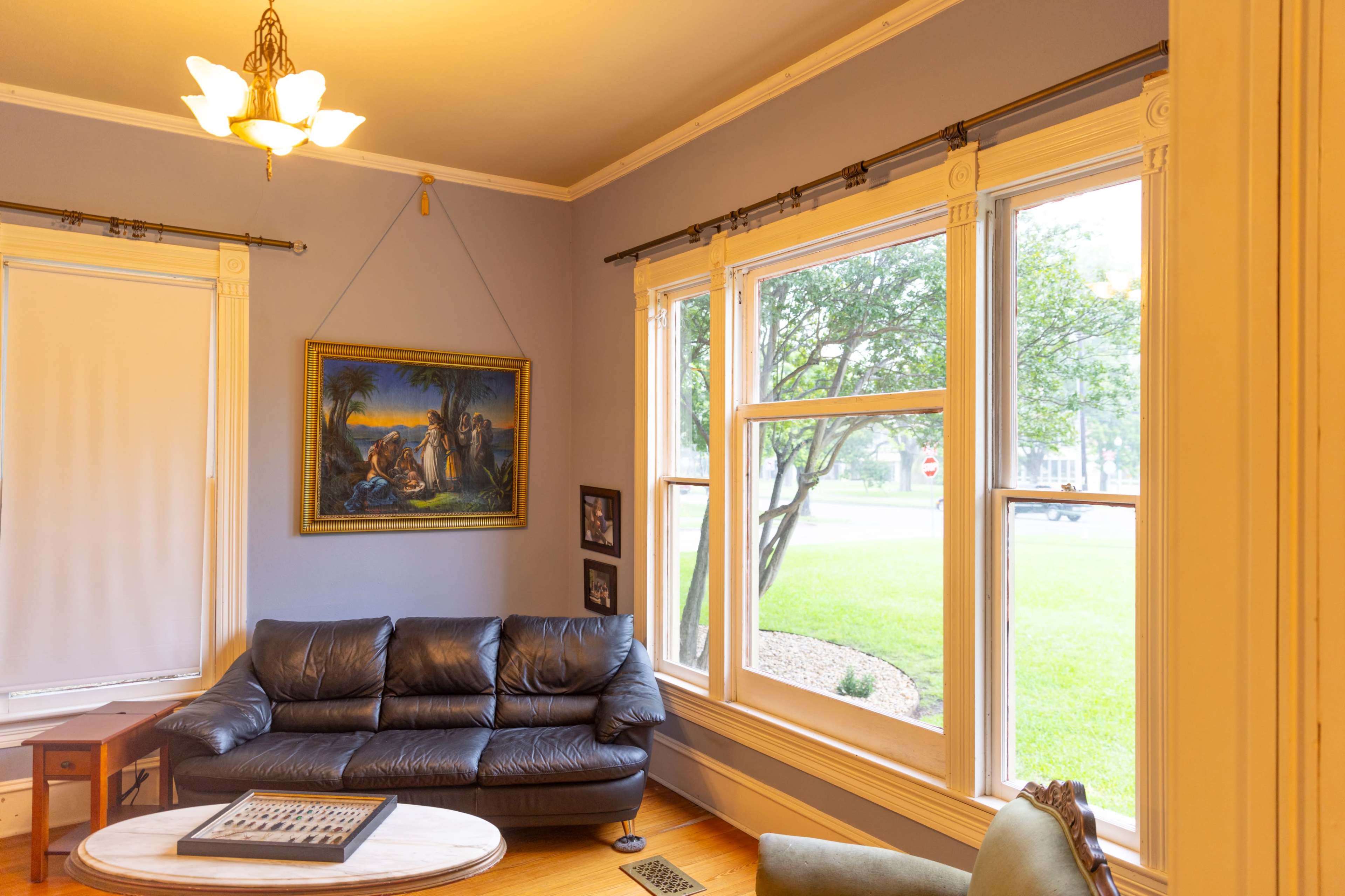 A living room with a leather sofa, a round coffee table, a framed painting on the wall, and large windows facing a green lawn.