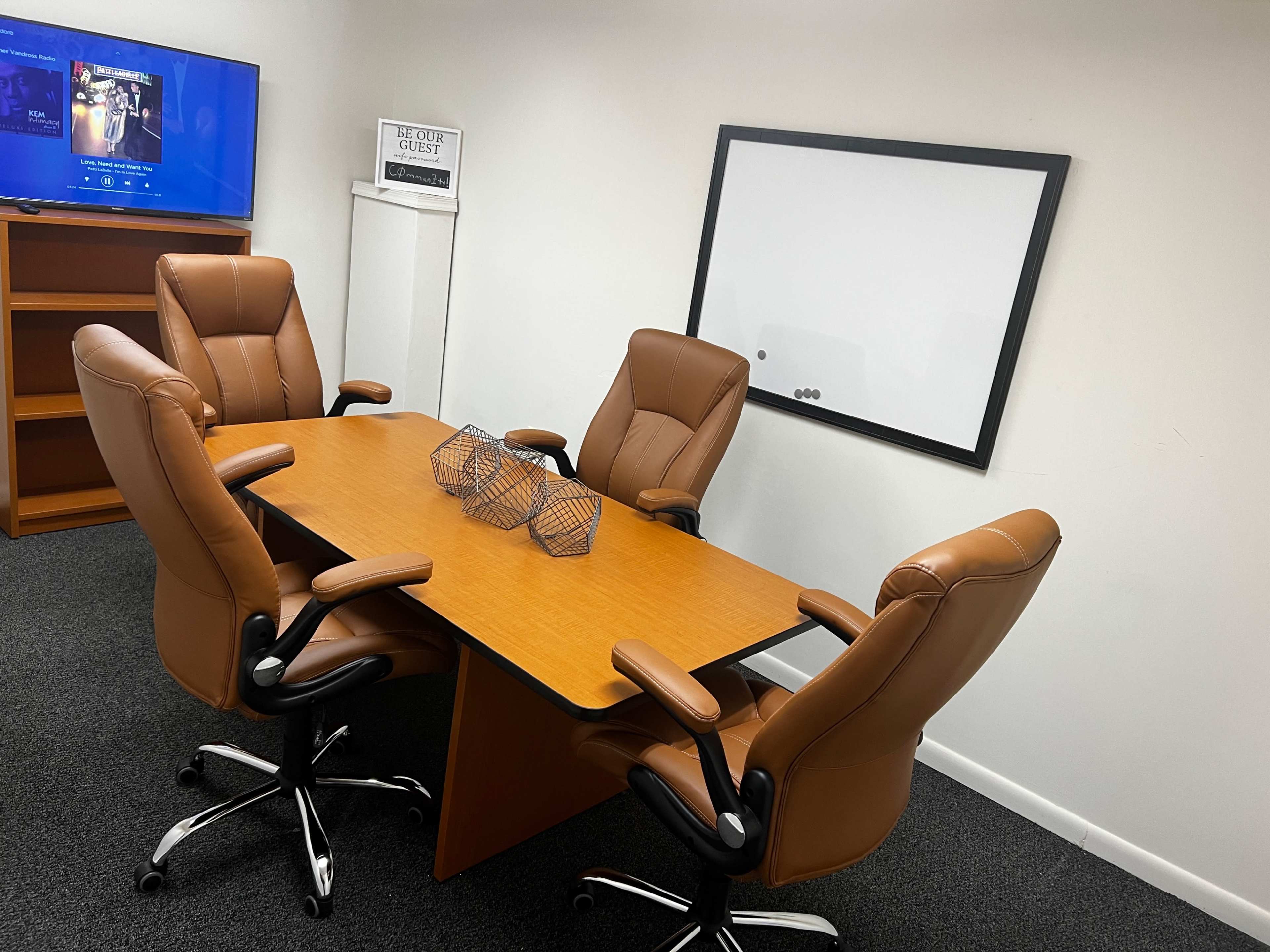 A conference room with a wooden table and four brown office chairs, along with a television on the wall and a whiteboard.