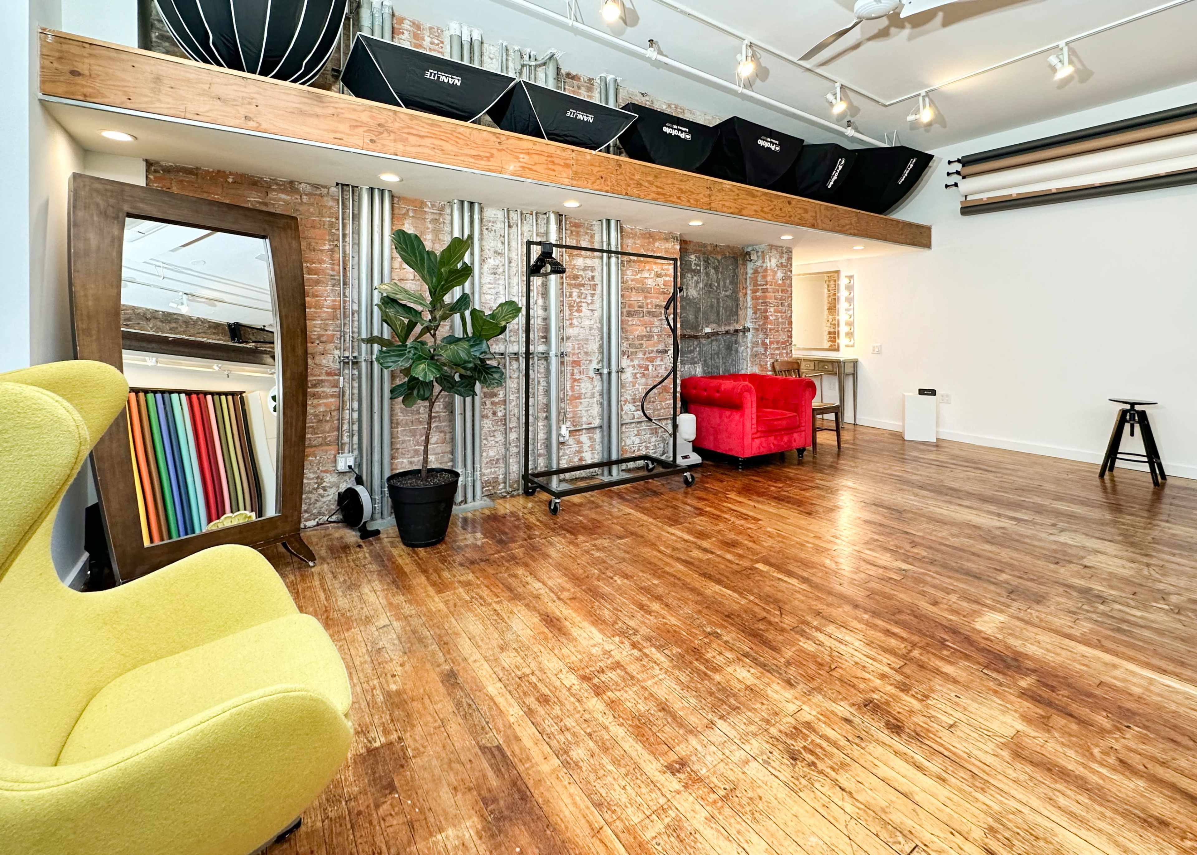 A photography studio with wooden floors, exposed brick walls, a large mirror, a yellow chair, a red chair, and various lighting equipment hanging above.