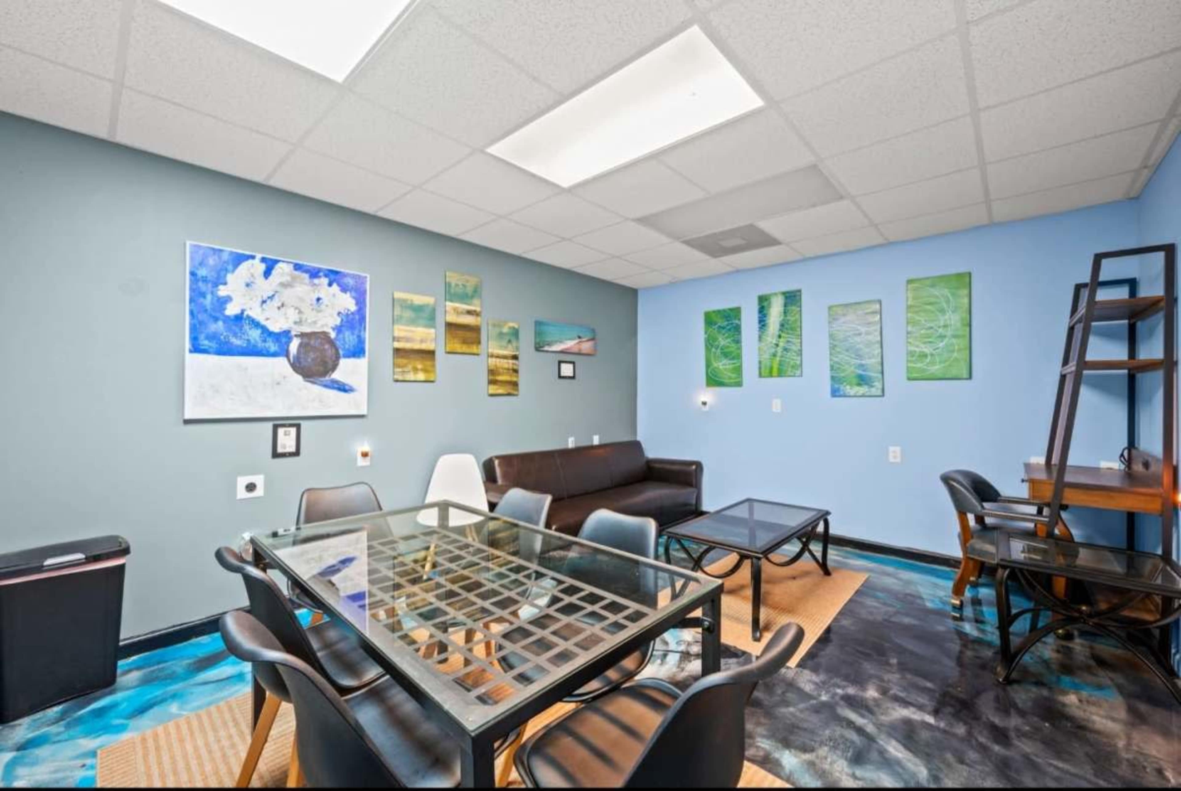 The image shows a modern lounge space equipped with a glass table, black chairs, and various abstract artworks on the walls.