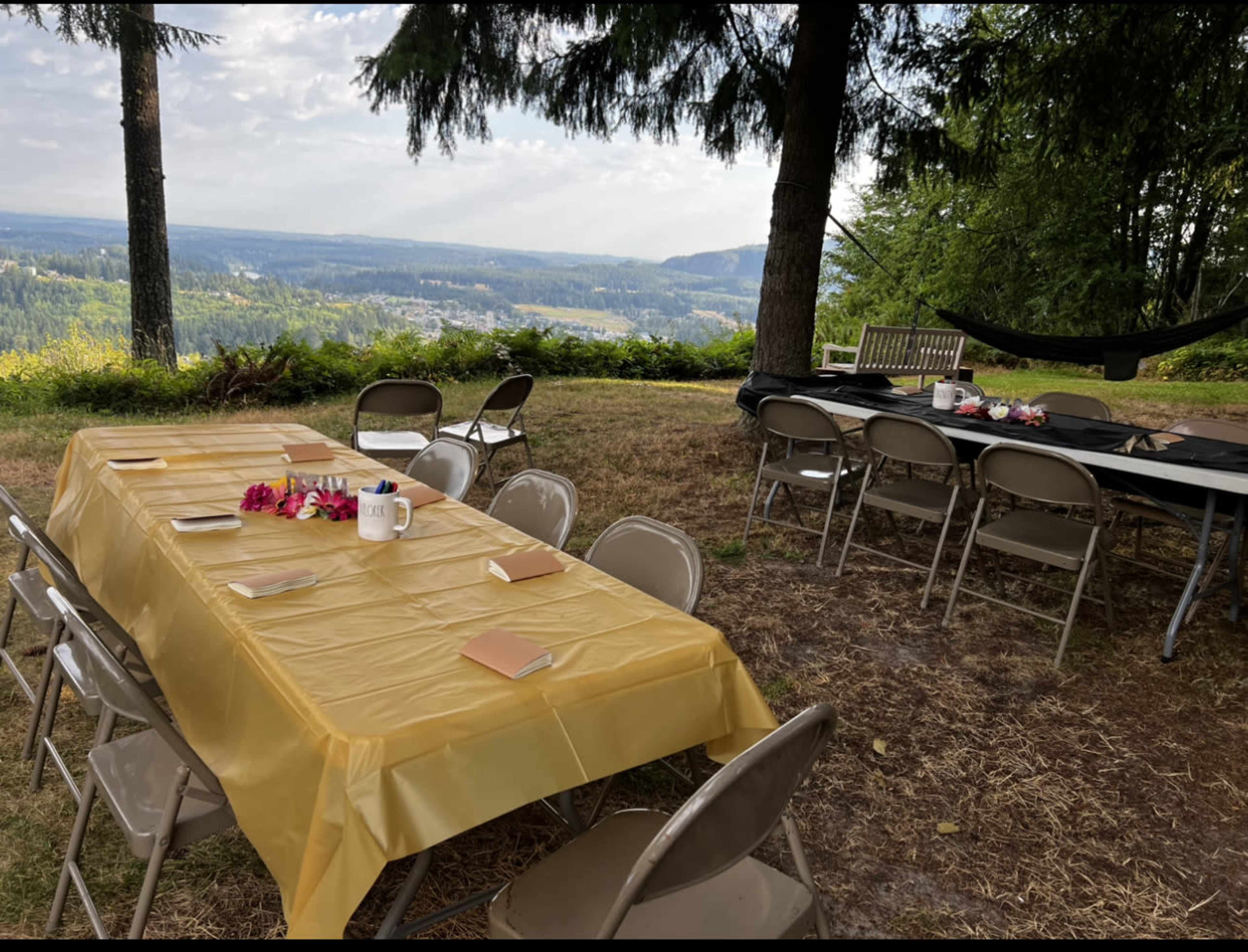 A long table covered with a yellow tablecloth is set outdoors on a grassy area, surrounded by chairs and offering a view of the distant landscape.