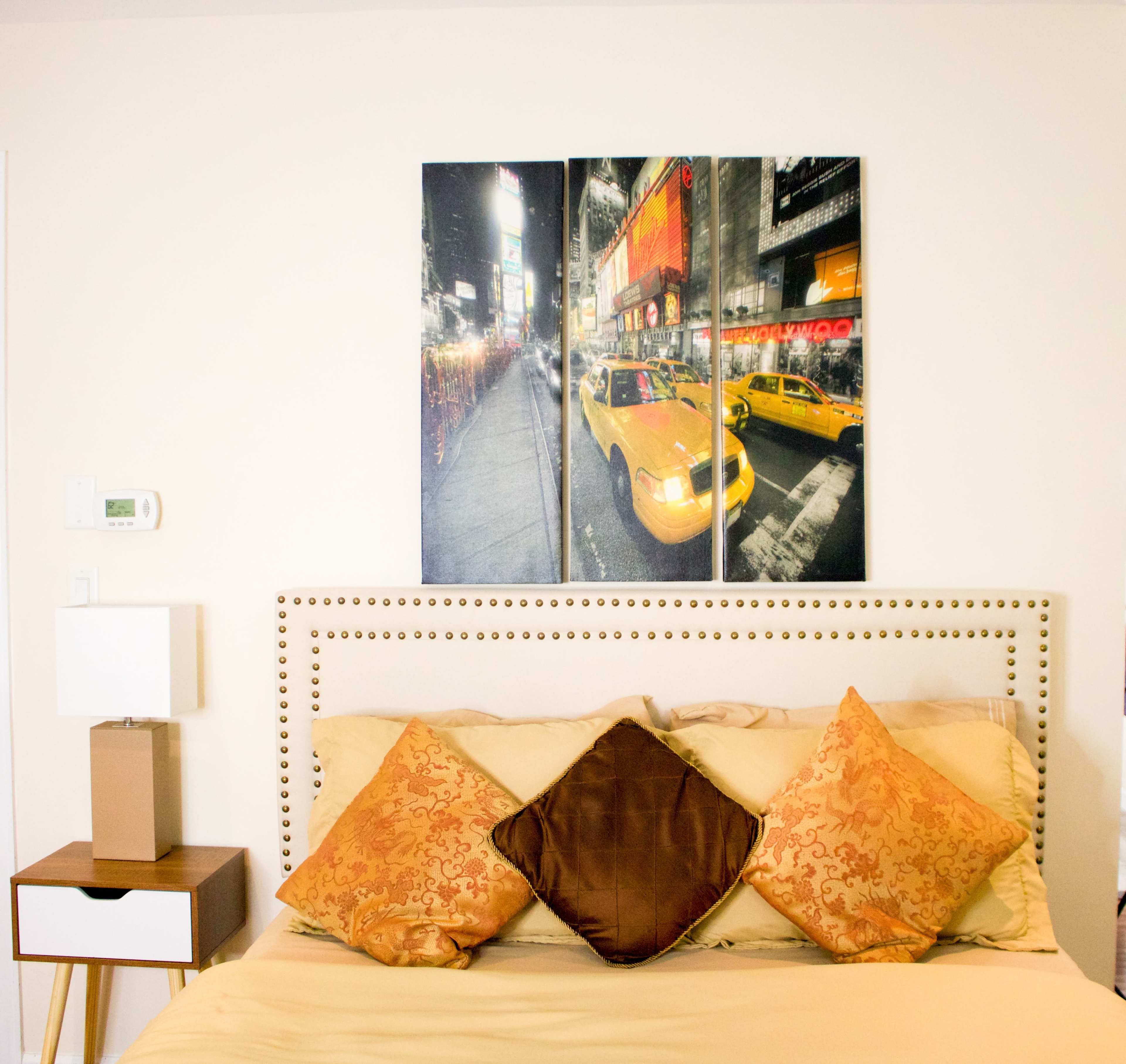 A bed with decorative pillows and a bedside table, featuring a large canvas artwork of yellow taxis in a city setting above the headboard.