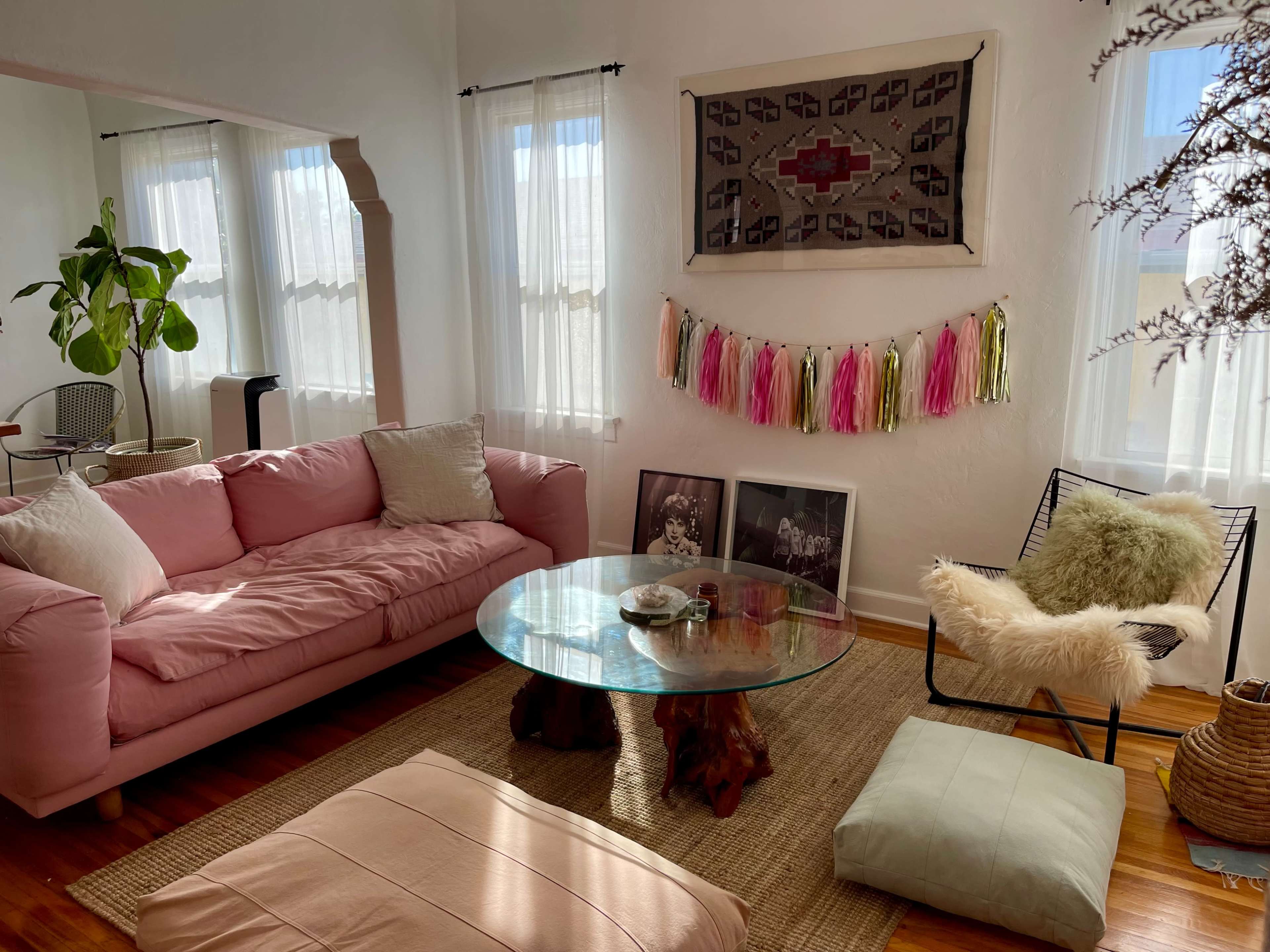 The image shows a bright living room with a pink sofa, a glass coffee table, framed artwork on the floor, and decorative elements like a wool throw and a plant.