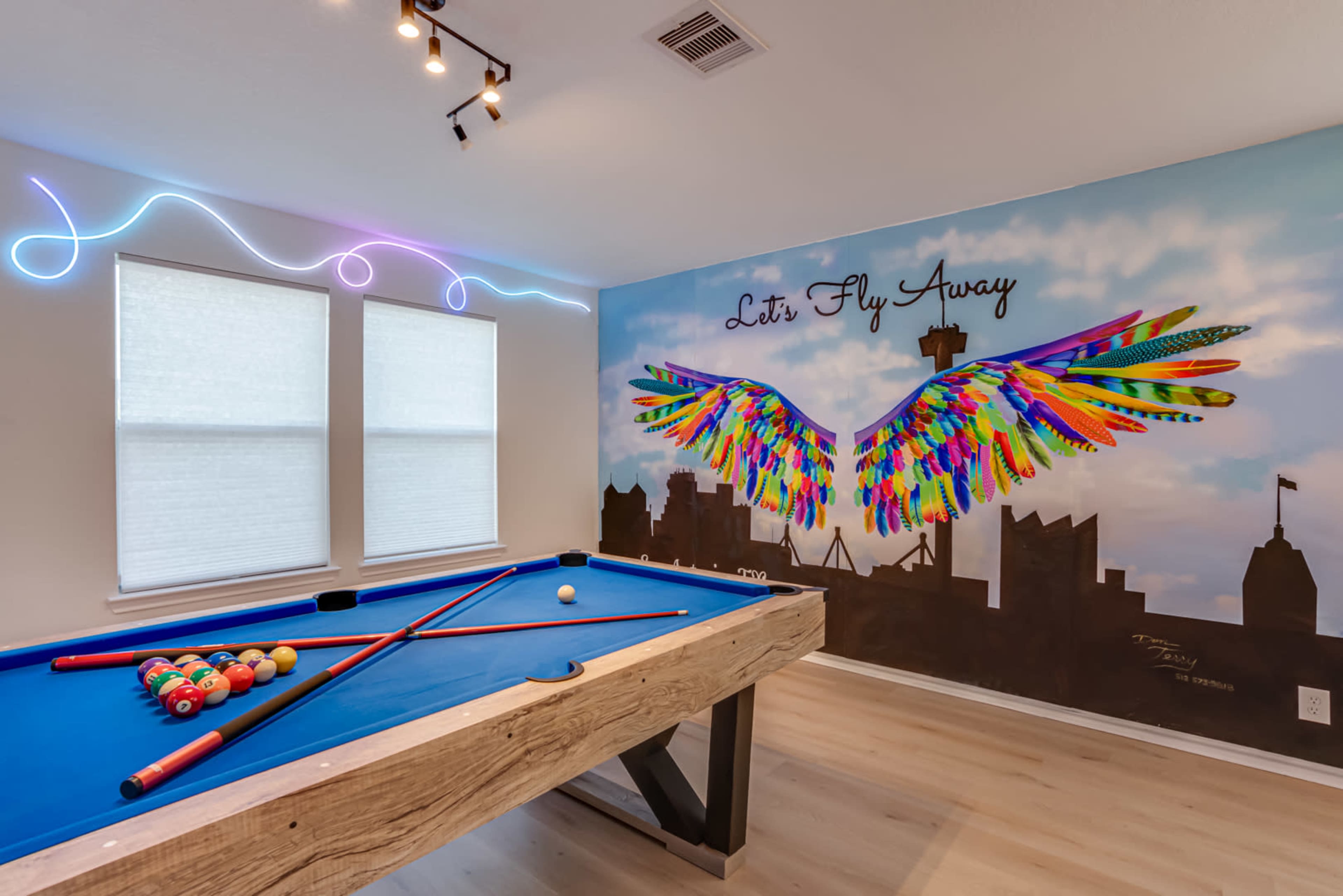A brightly colored game room featuring a blue pool table in front of a mural with vibrant wings and the phrase "Let's Fly Away" on the wall.