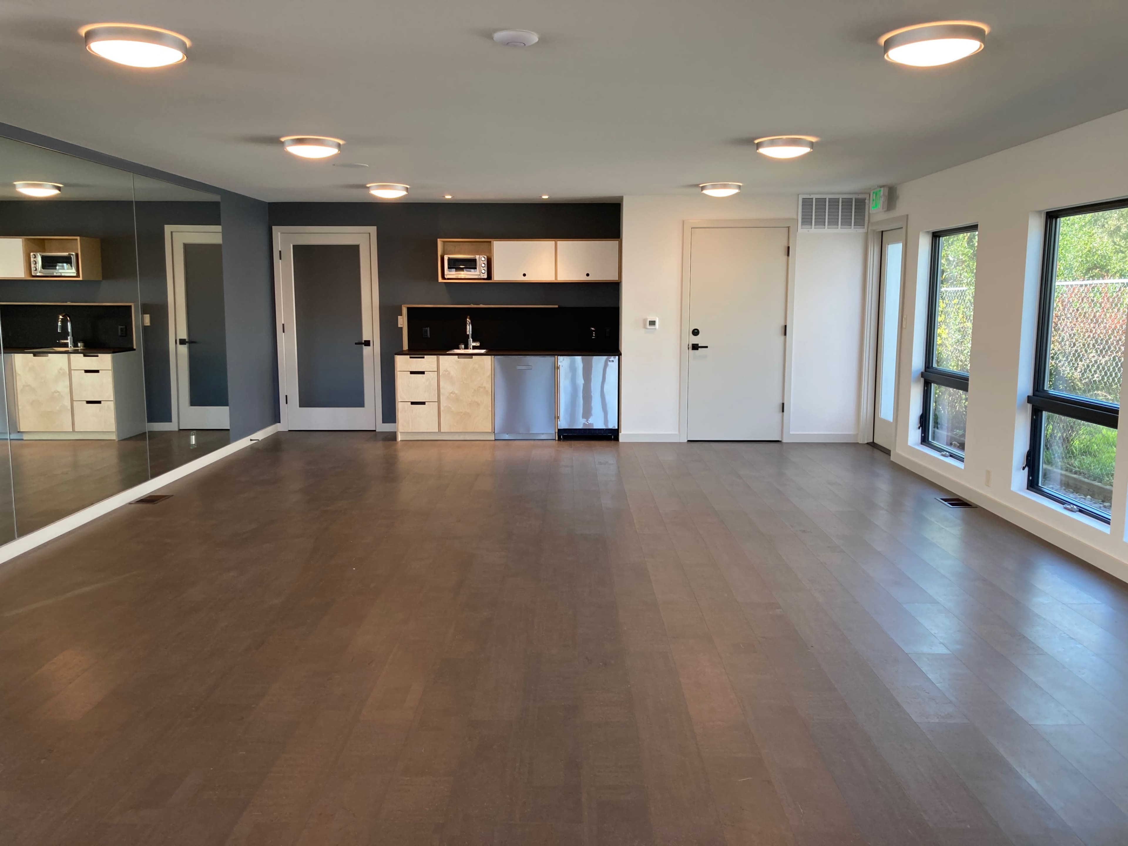 A spacious, modern room with a mirrored wall, light fixtures on the ceiling, a small kitchenette in the corner, and large windows providing natural light.