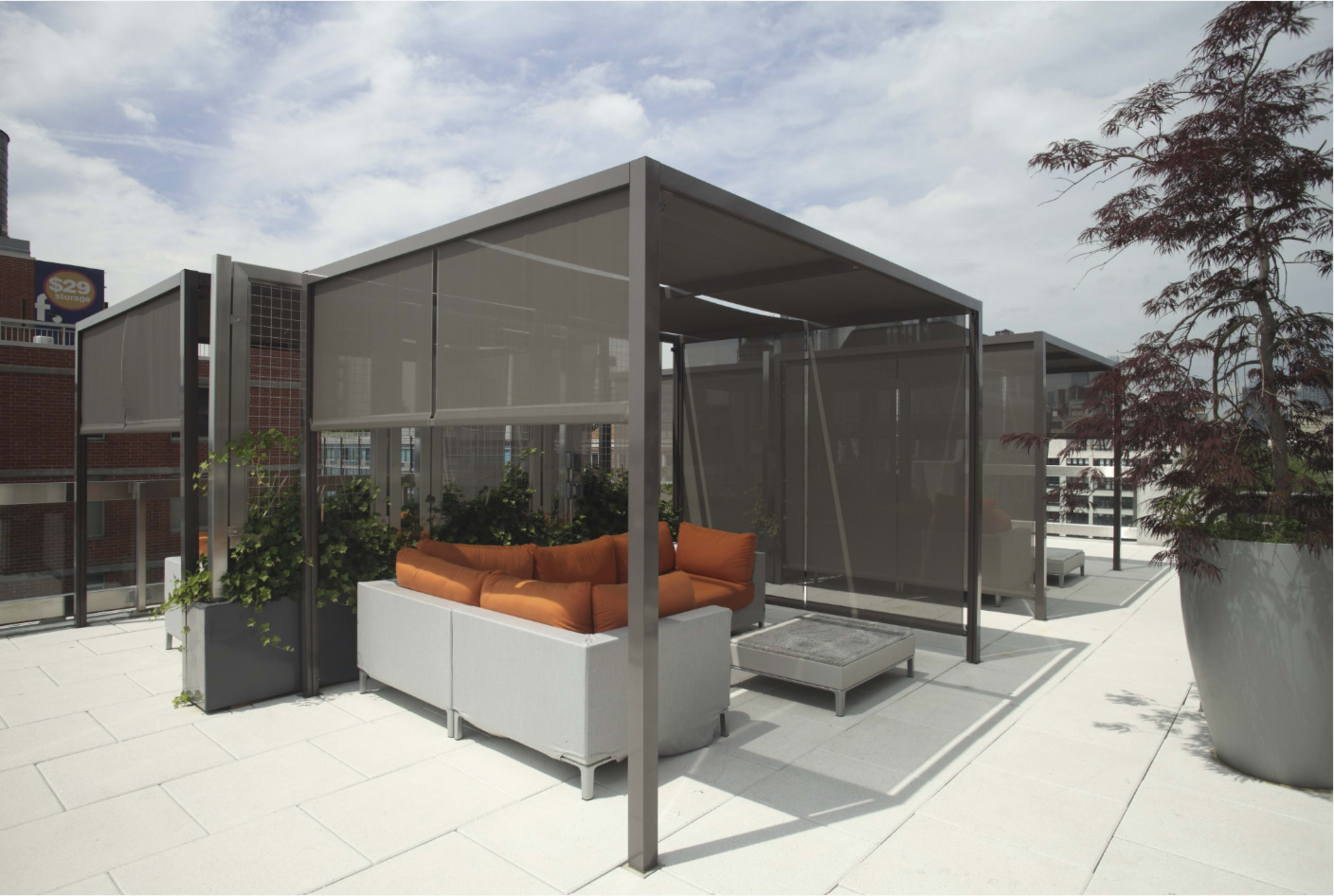 The image shows a modern rooftop lounge area with a gray sofa and orange cushions, surrounded by mesh screens and potted plants.