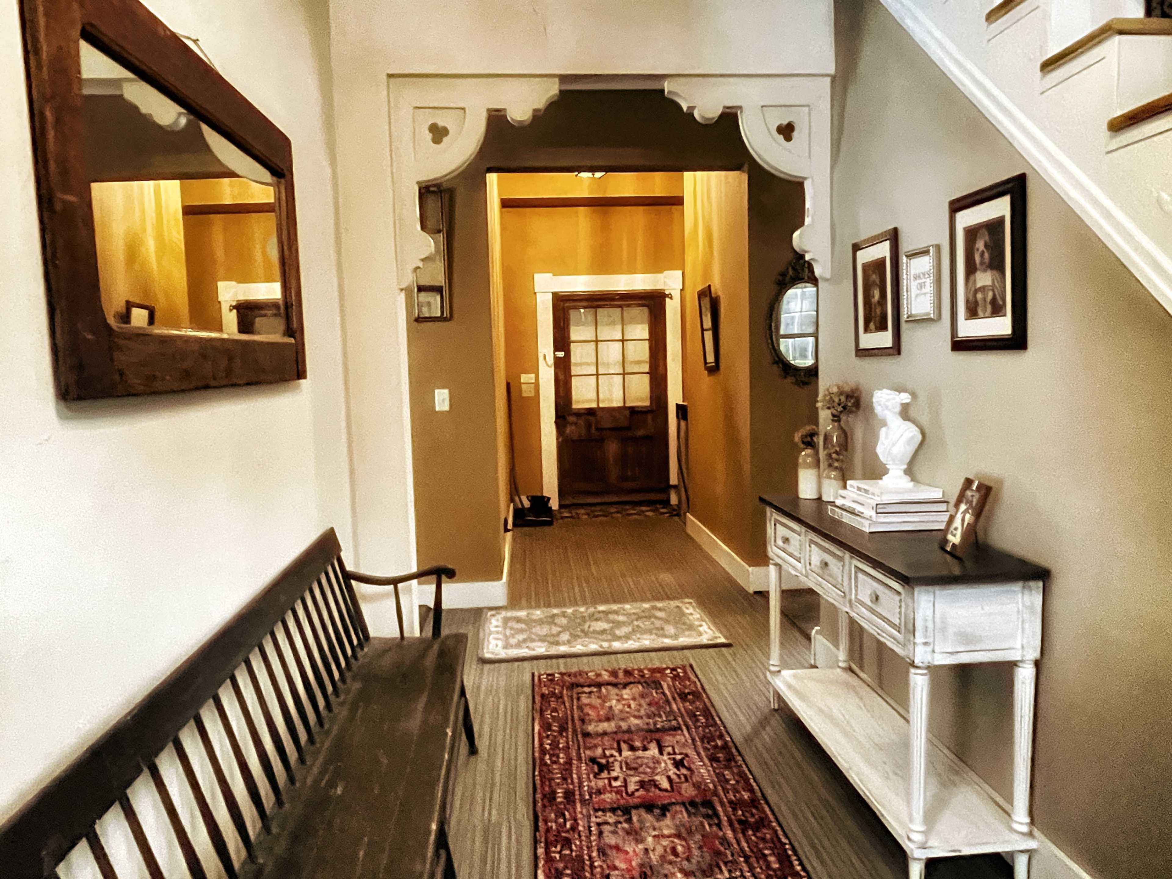 A hallway with a wooden bench, a side table displaying framed photos, and a staircase leading to an upper level.