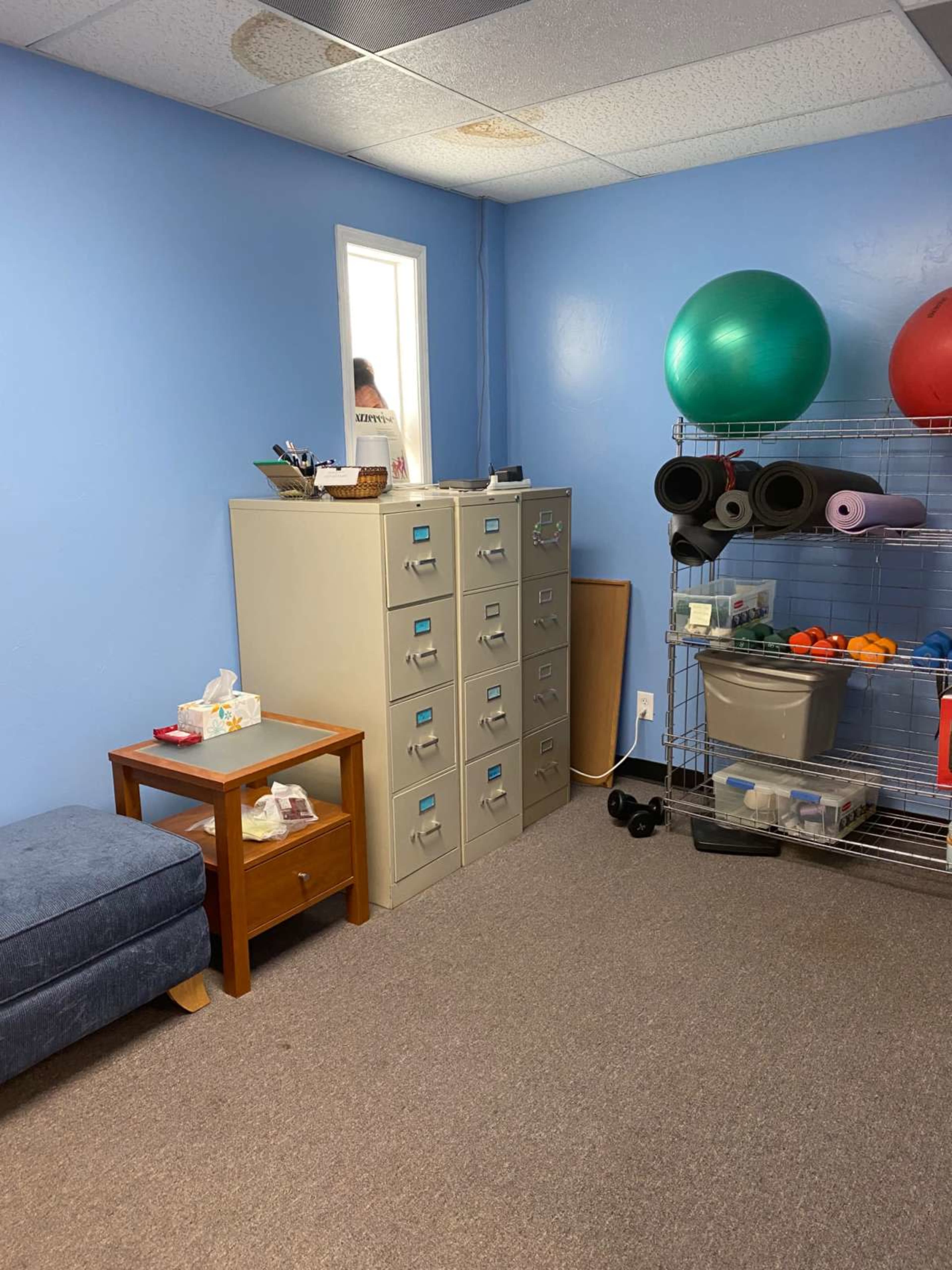 The room features a metal filing cabinet, a wooden side table, exercise equipment, and fitness balls against a blue wall.