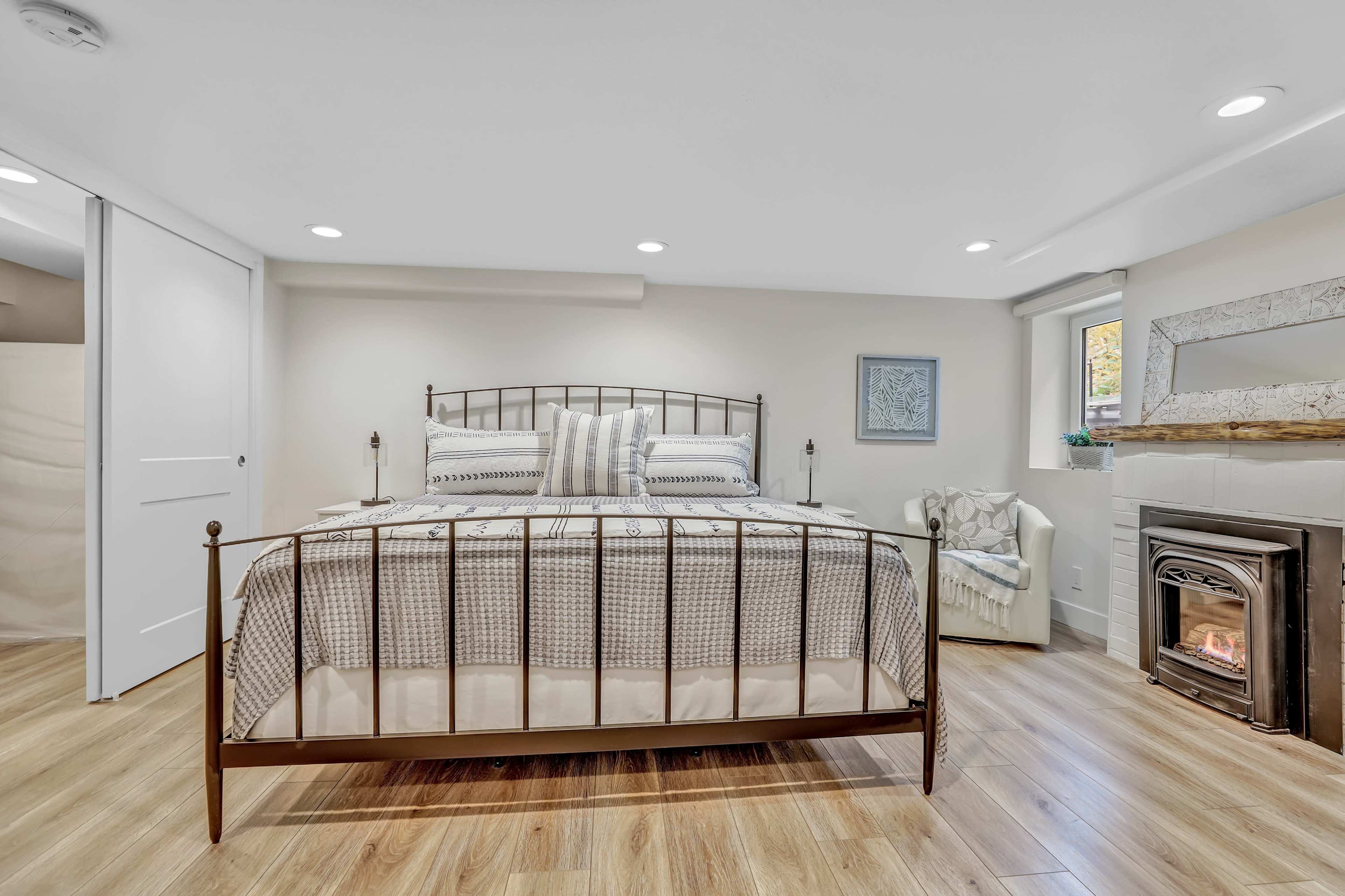 A neatly arranged bedroom features a metal bed frame with a patterned duvet, a cozy chair in the corner, and a small fireplace.