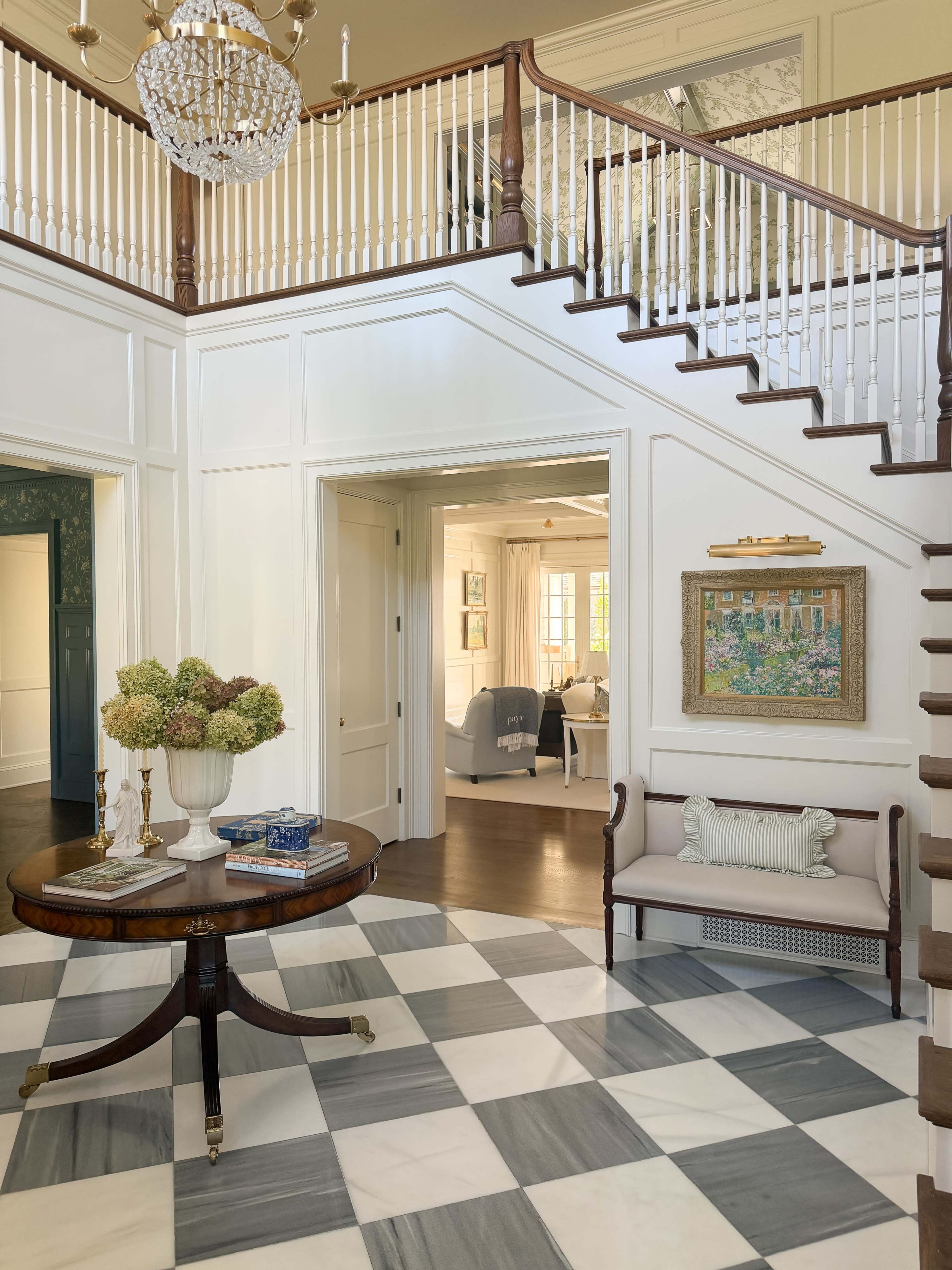 The image shows a well-lit entryway featuring a decorative round table and a staircase with a wooden railing, set against a backdrop of marble checkered flooring.