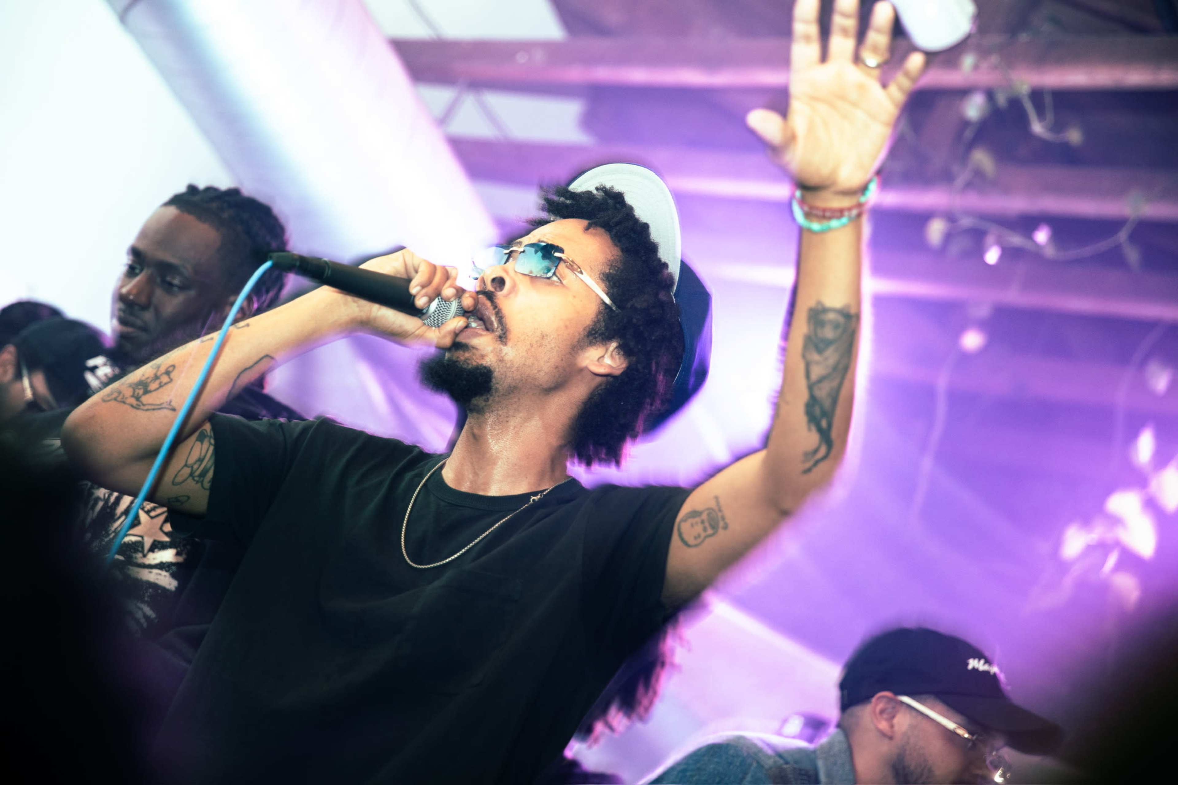A man with curly hair and sunglasses performs on stage with a microphone while others are visible in the background.