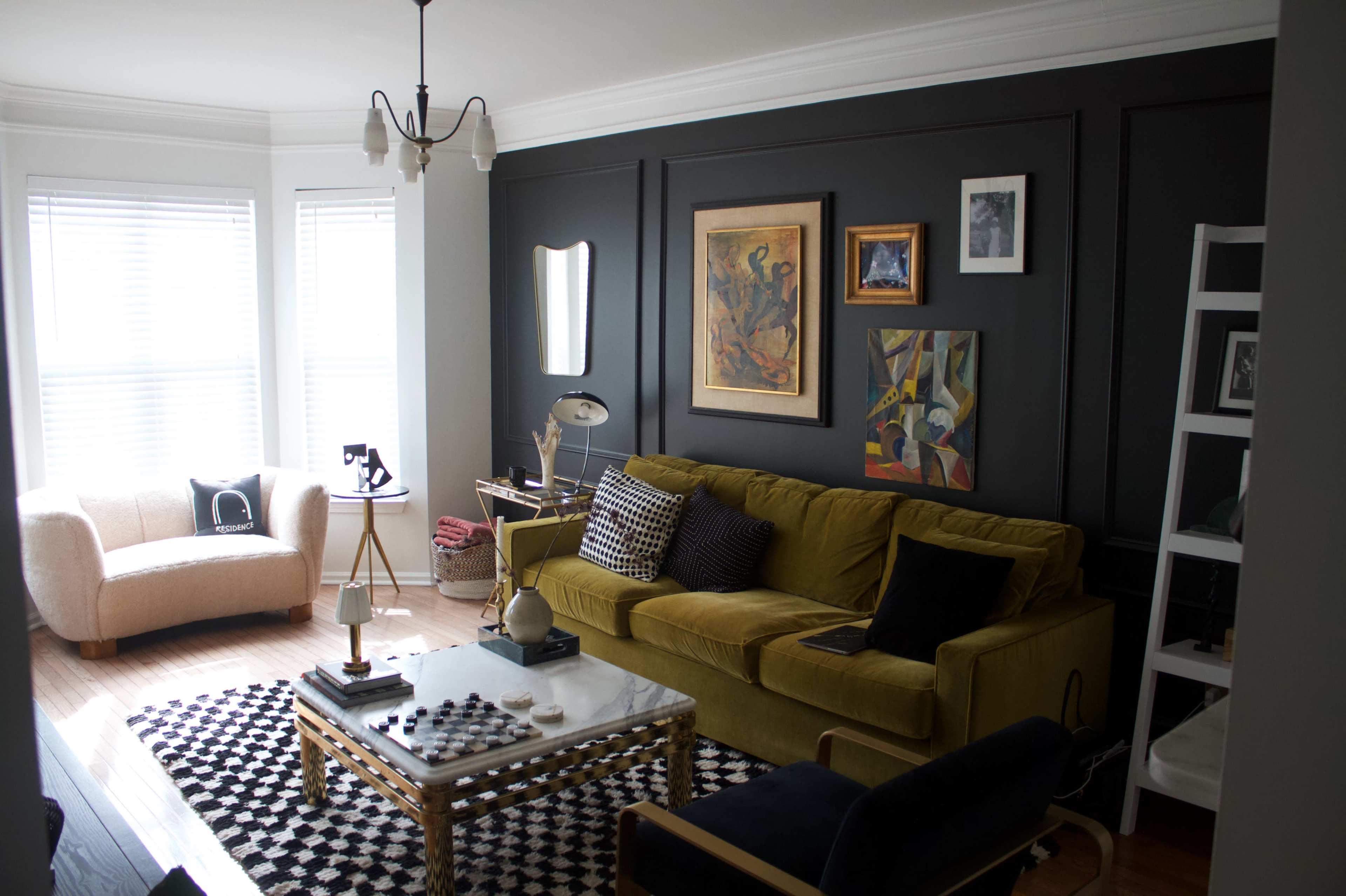 A living room featuring a green sofa, a coffee table, artwork on the walls, and large windows with white blinds.