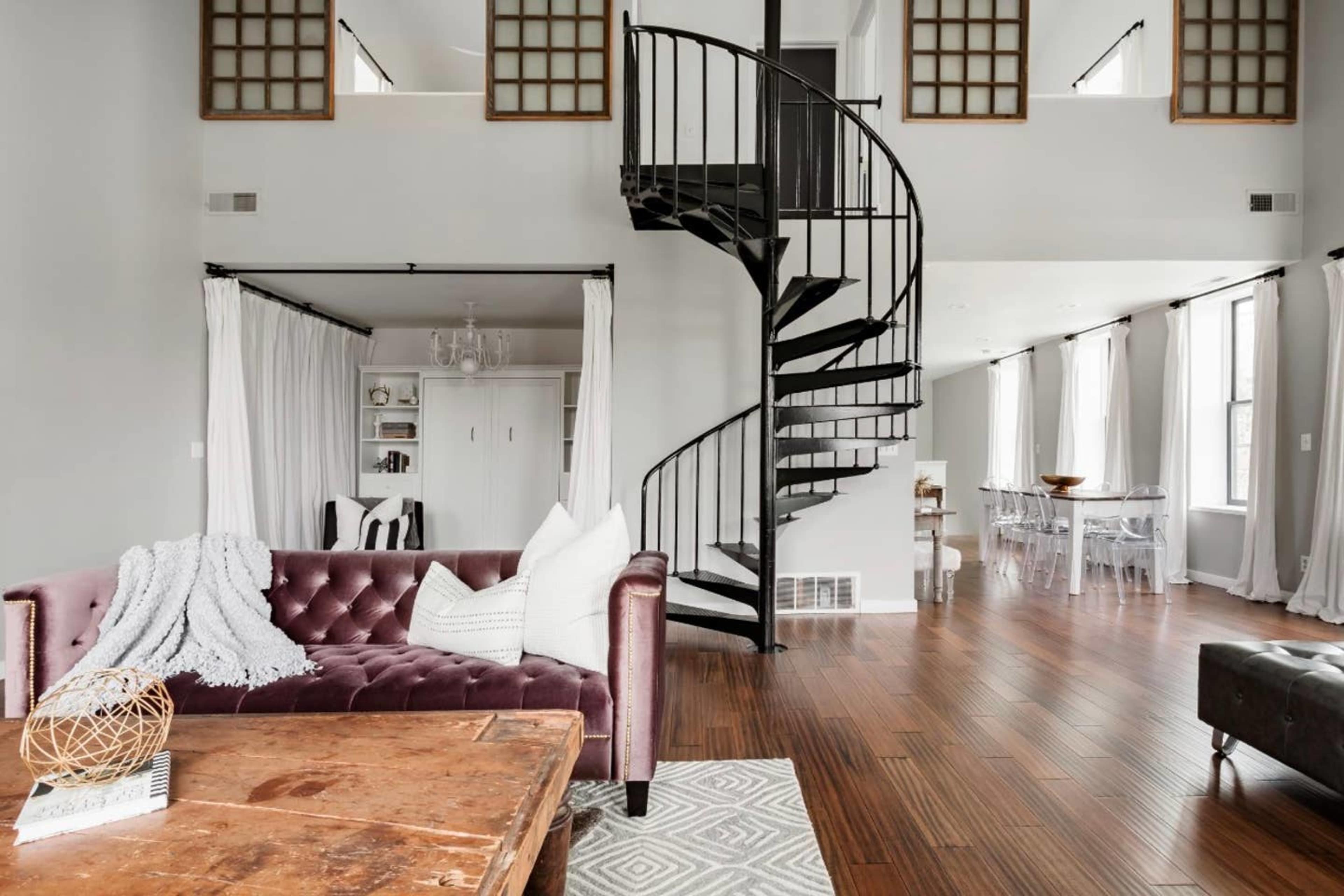 A modern living space with a spiral staircase, a velvet sofa, wooden flooring, and large windows allowing natural light.