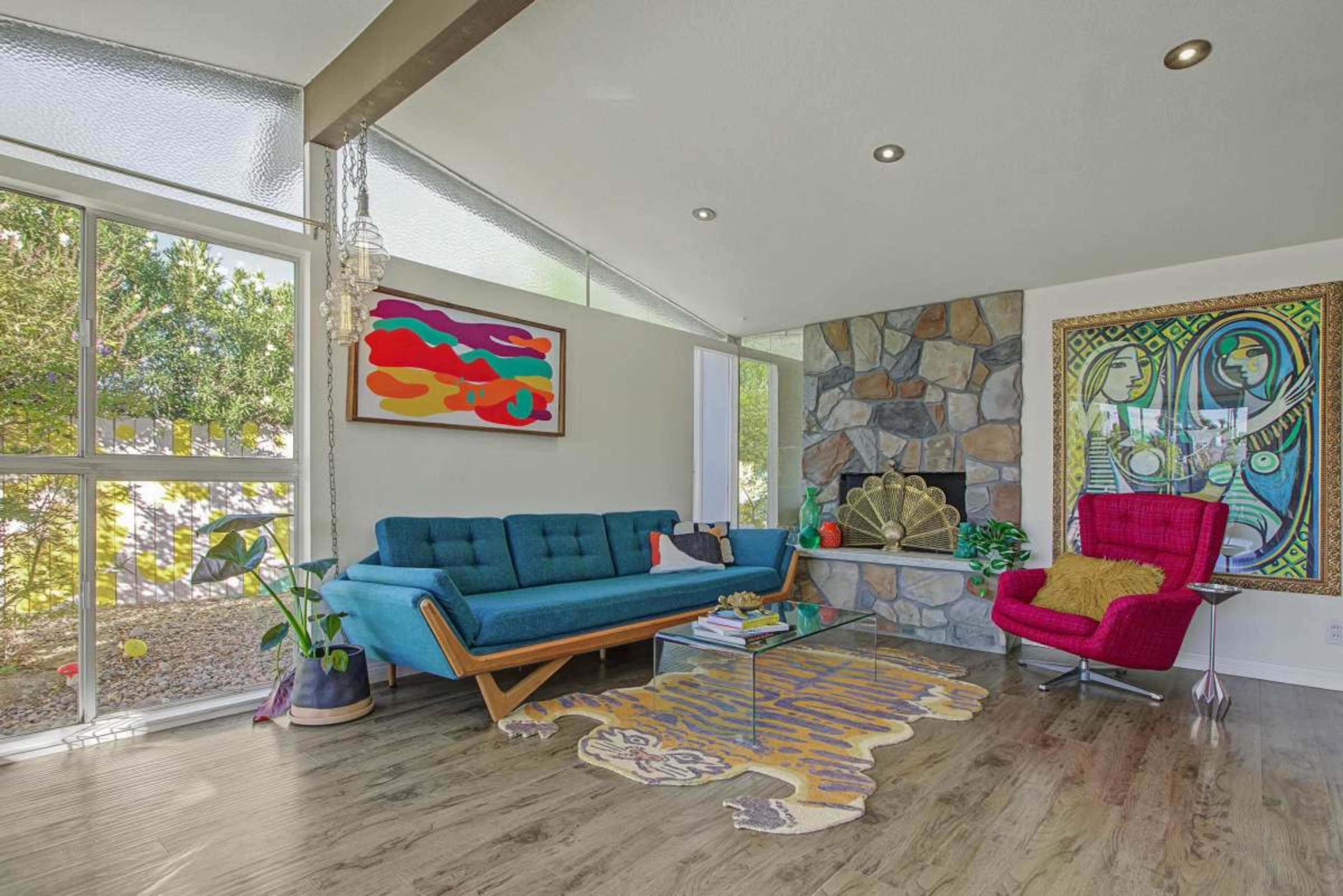 The living room features a blue mid-century modern sofa, a pink armchair, a stone fireplace, and colorful artwork on the walls.