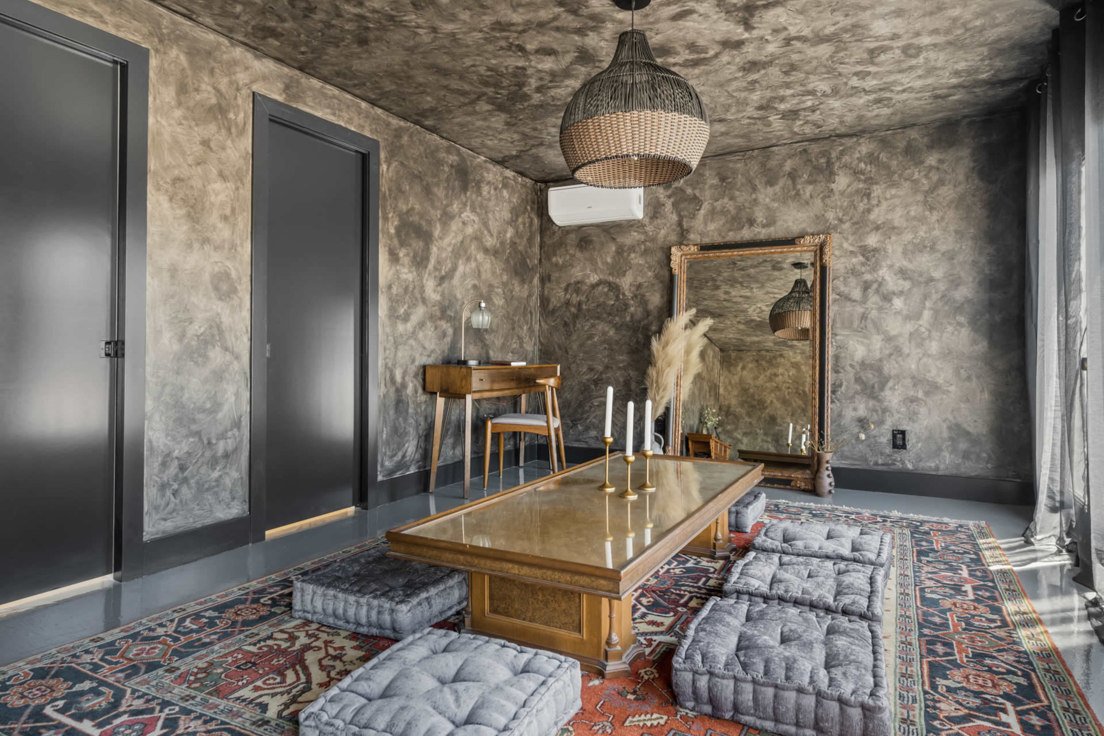 The image shows a minimalist room with a textured ceiling, a central wooden table surrounded by cushioned seats, and a large mirror against a wall.