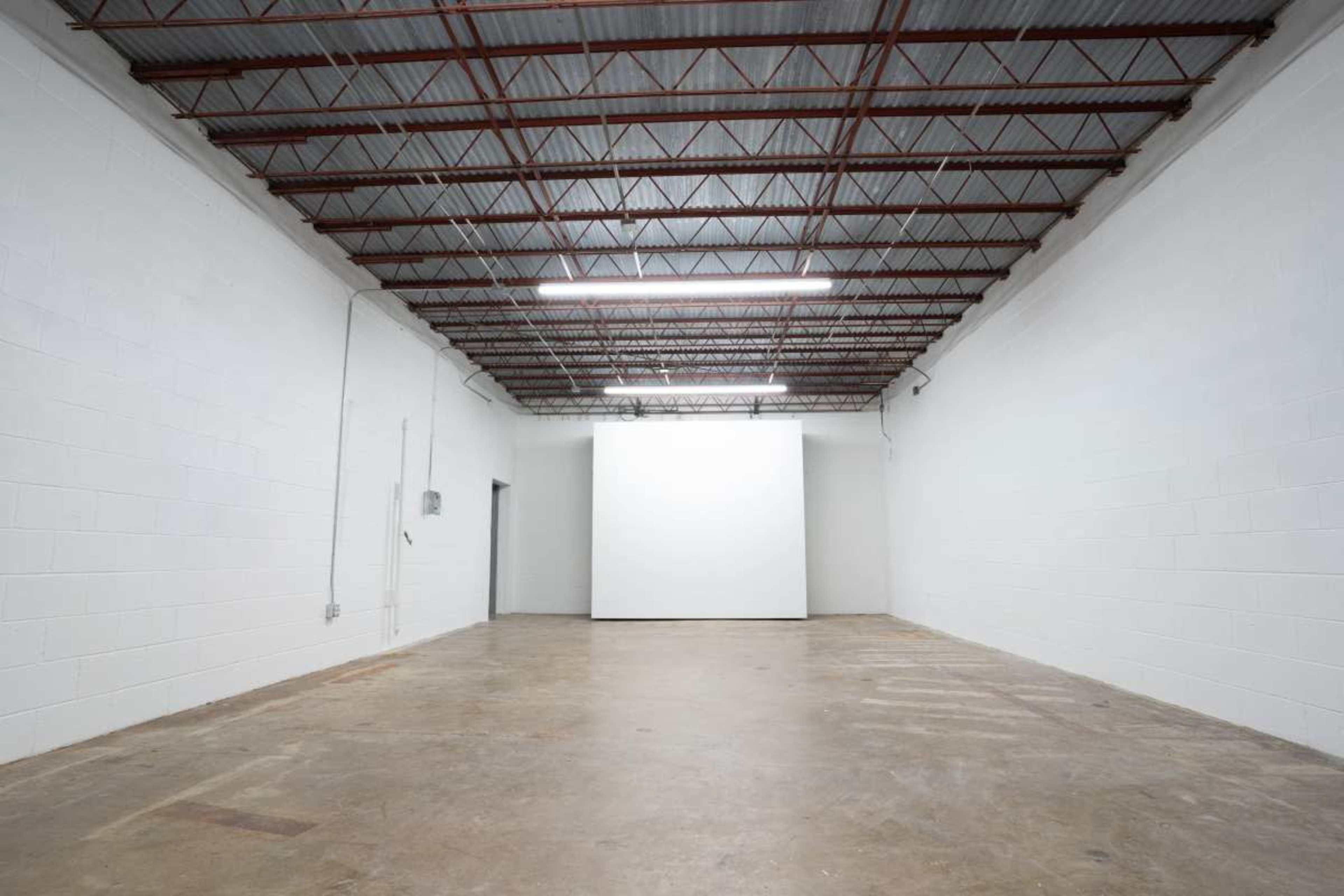 The image shows a large, empty room with white walls, a concrete floor, and a blank wall at the far end.