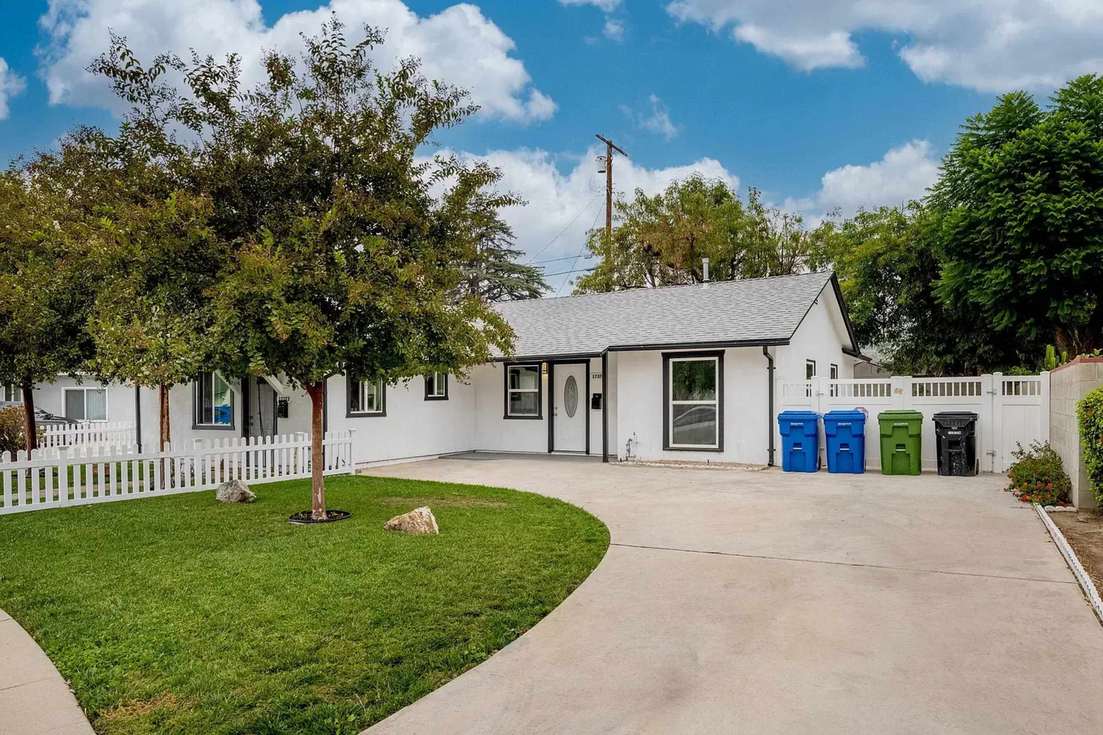 A single-story house with a gray roof and white exterior is surrounded by a green lawn and features a curved driveway with three colored trash bins.