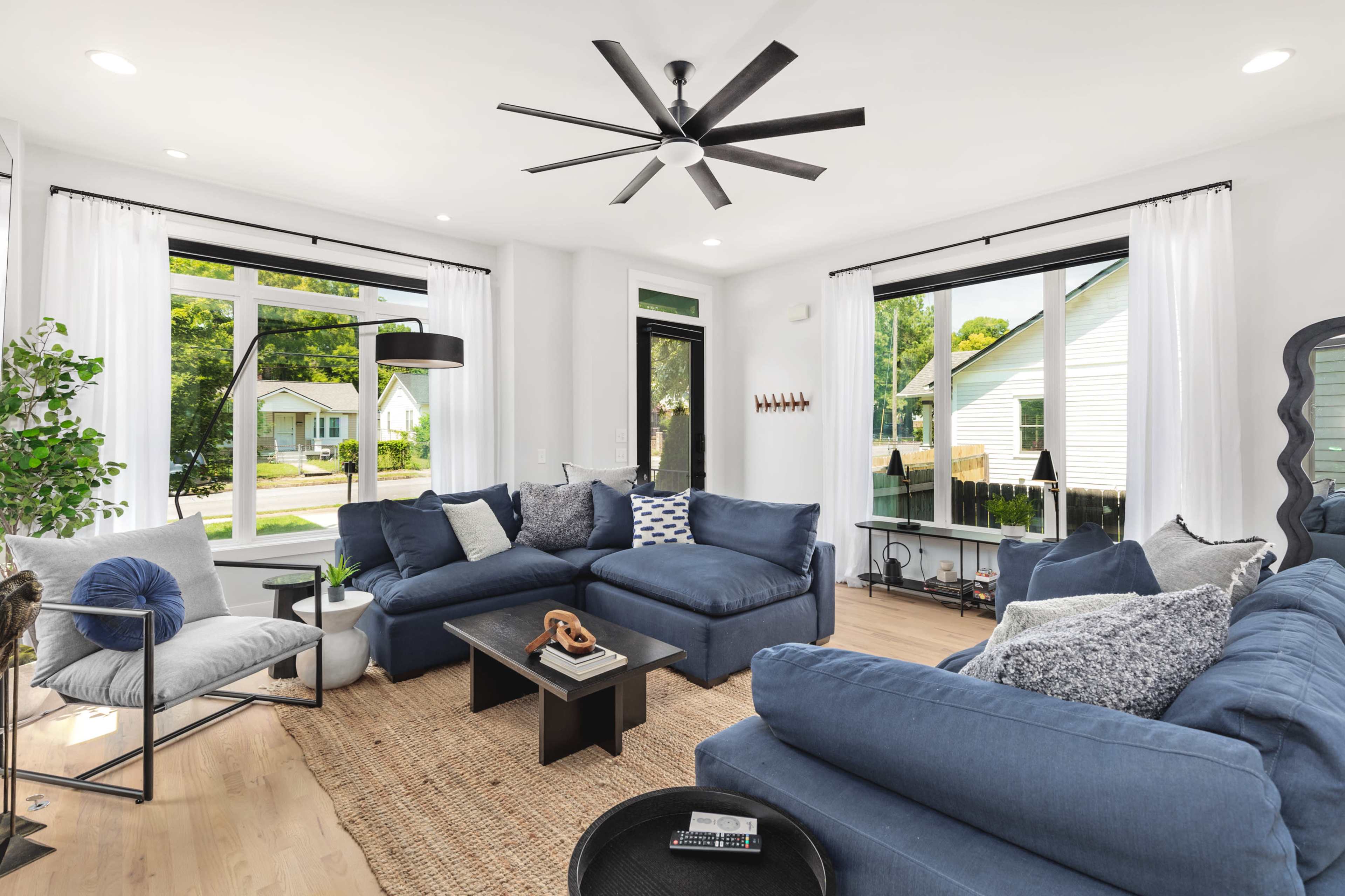 A bright living room features blue sectional sofas, a coffee table, and large windows with sheer white curtains.