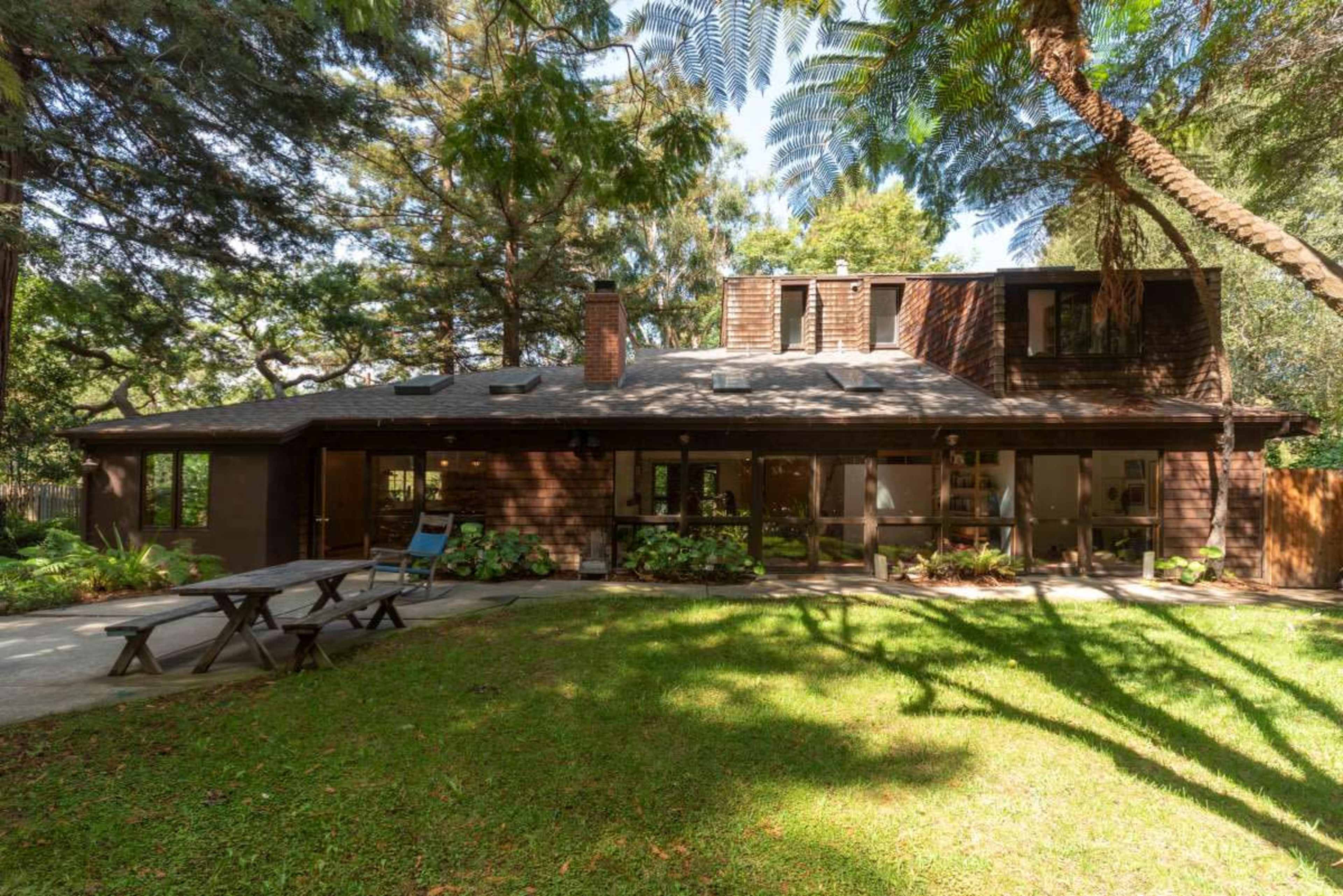 A wooden house with a sloped roof, surrounded by trees and a grassy area, features a patio and a picnic table in the backyard.