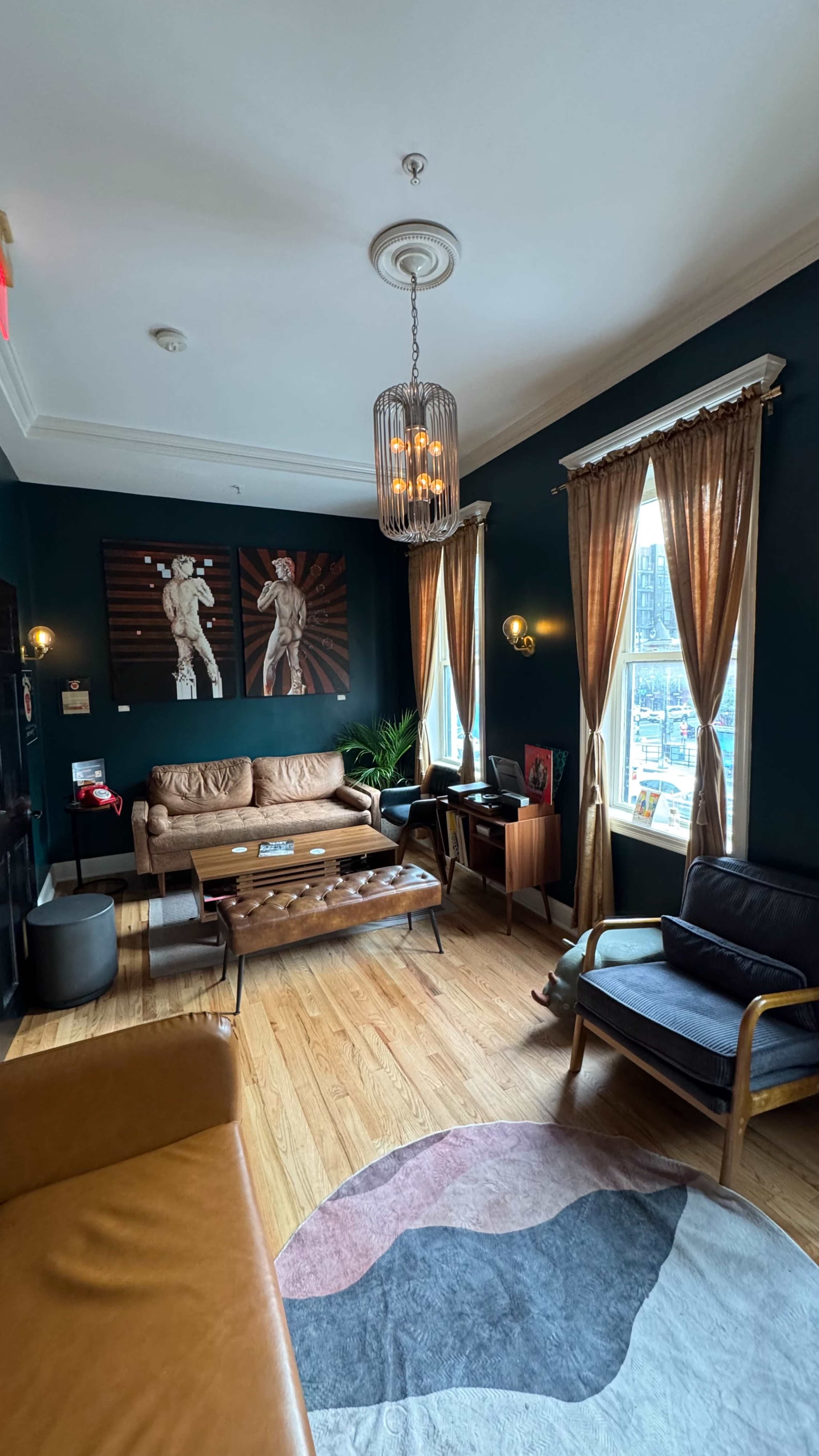 The image shows a stylish living room with dark teal walls, wooden flooring, a large window with curtains, and a mix of modern furniture including sofas and a coffee table.