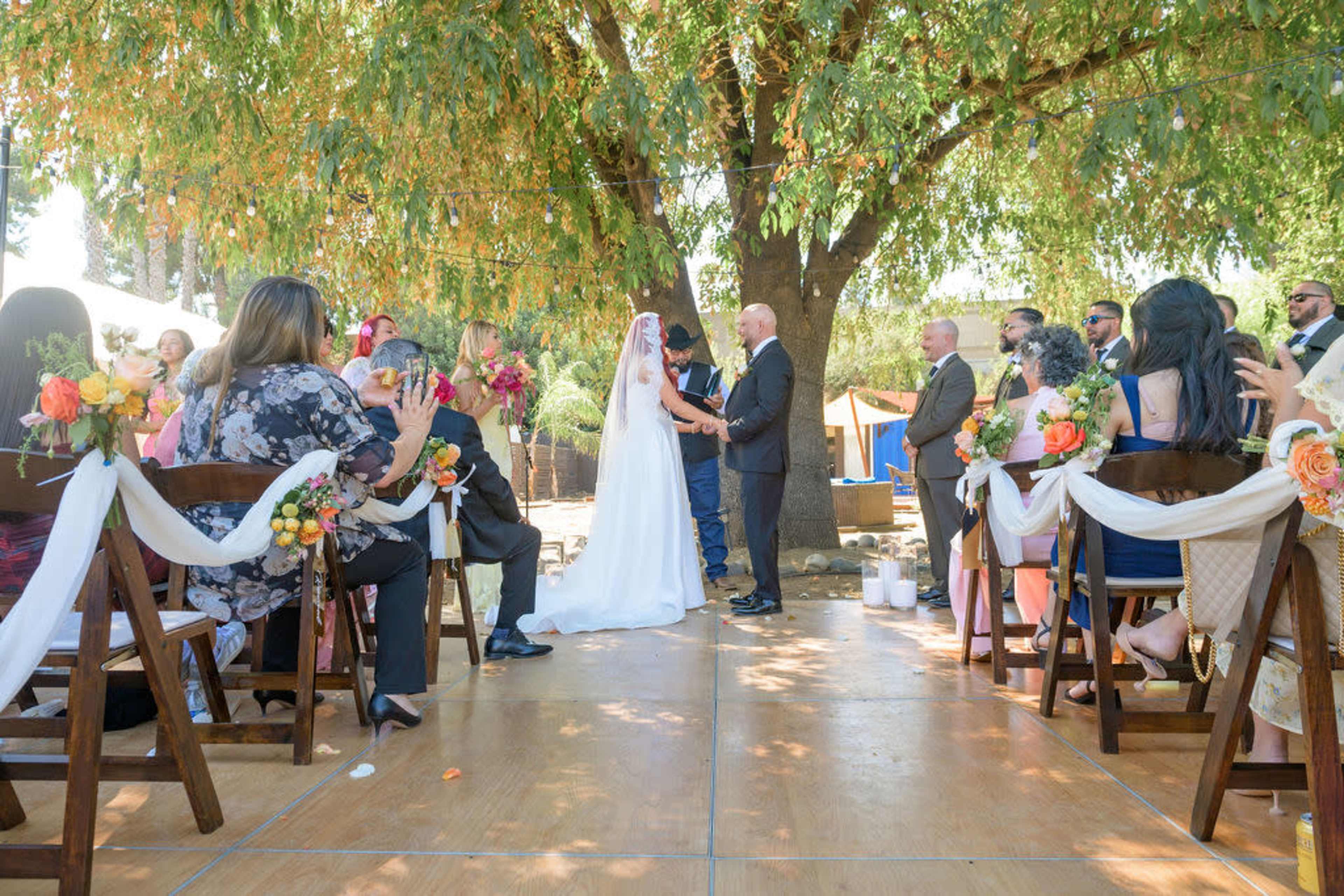 A couple stands under a large tree while exchanging vows at an outdoor wedding ceremony, surrounded by seated guests.