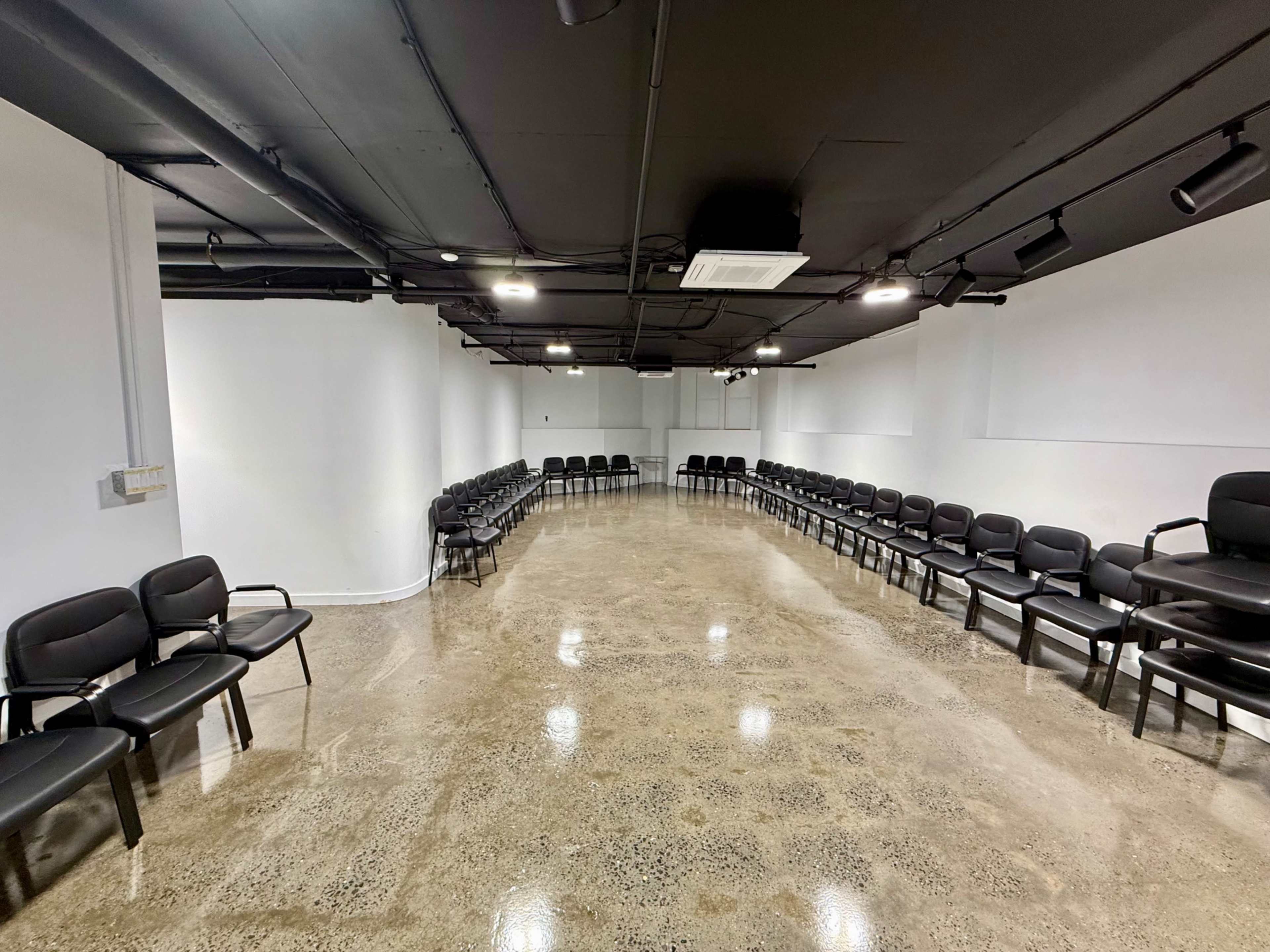 The image shows a large, empty room with a polished concrete floor and rows of black chairs arranged in a circular formation.