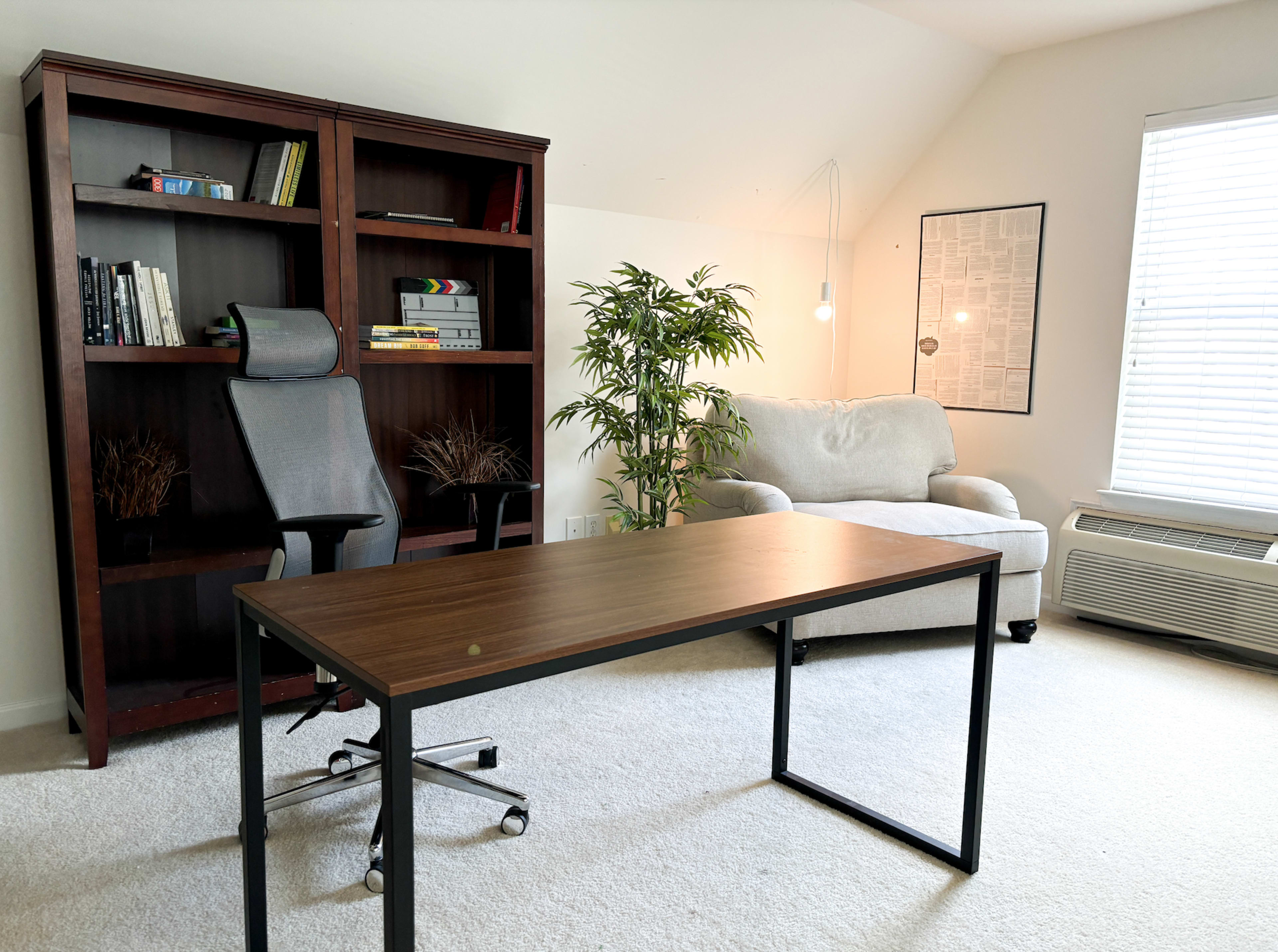A minimalist office space featuring a wooden desk, an ergonomic chair, a bookshelf filled with books, a potted plant, and a cozy couch beside a window.