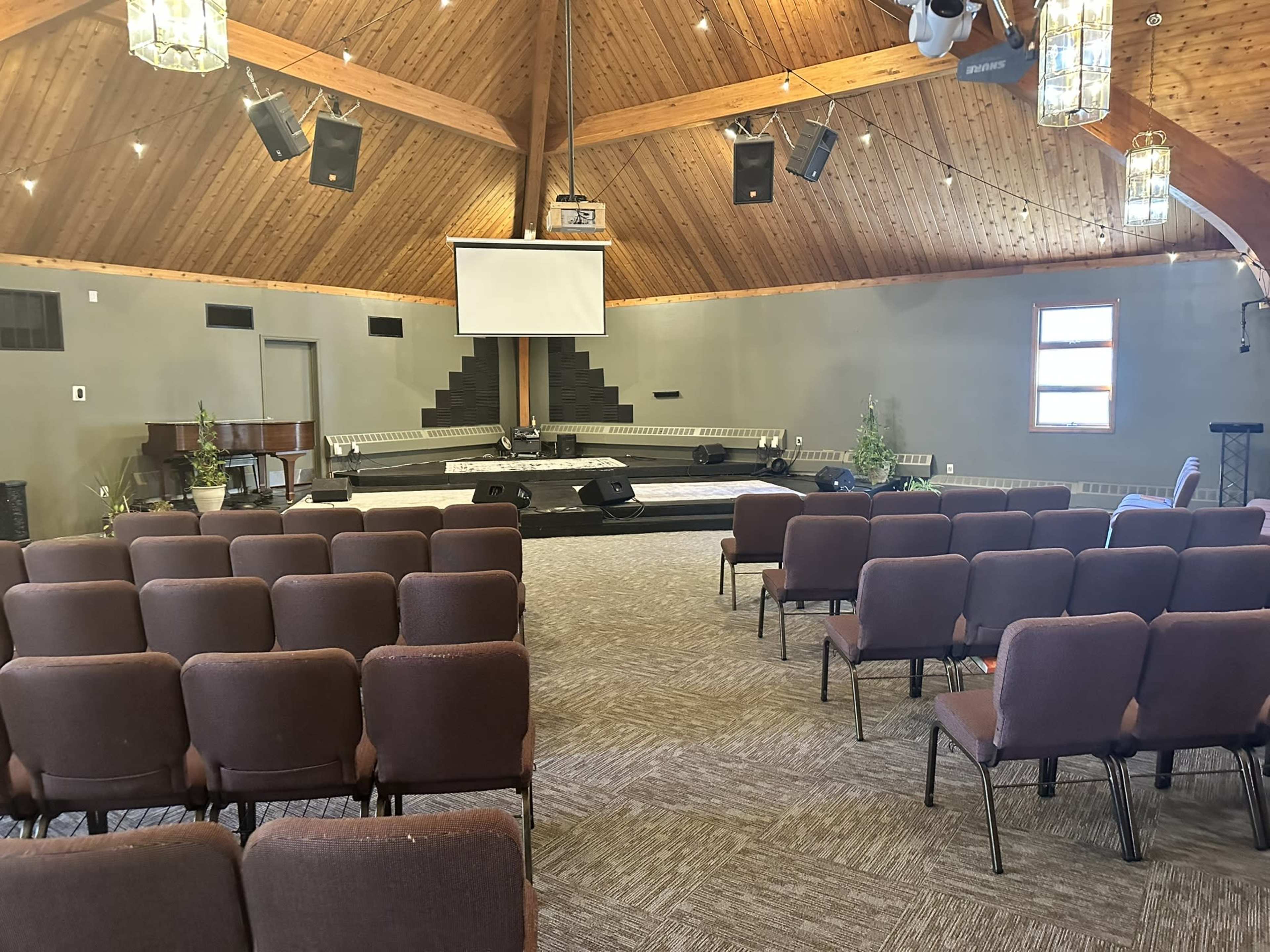 An interior of a spacious auditorium with rows of brown chairs facing a raised stage that features a projector screen and audio equipment.