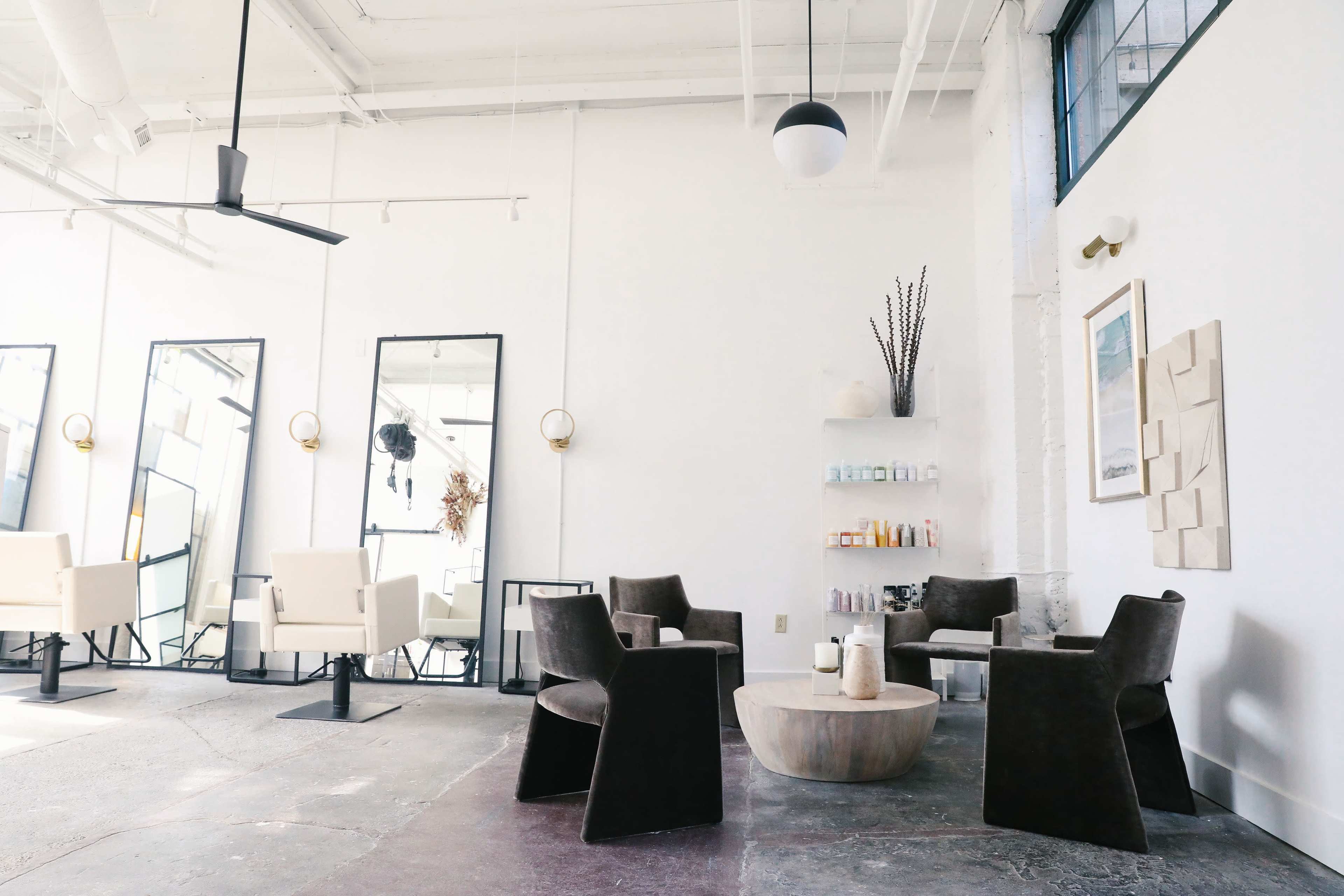 A modern salon interior featuring seating arrangements, mirrors, and a display shelf against a white wall.