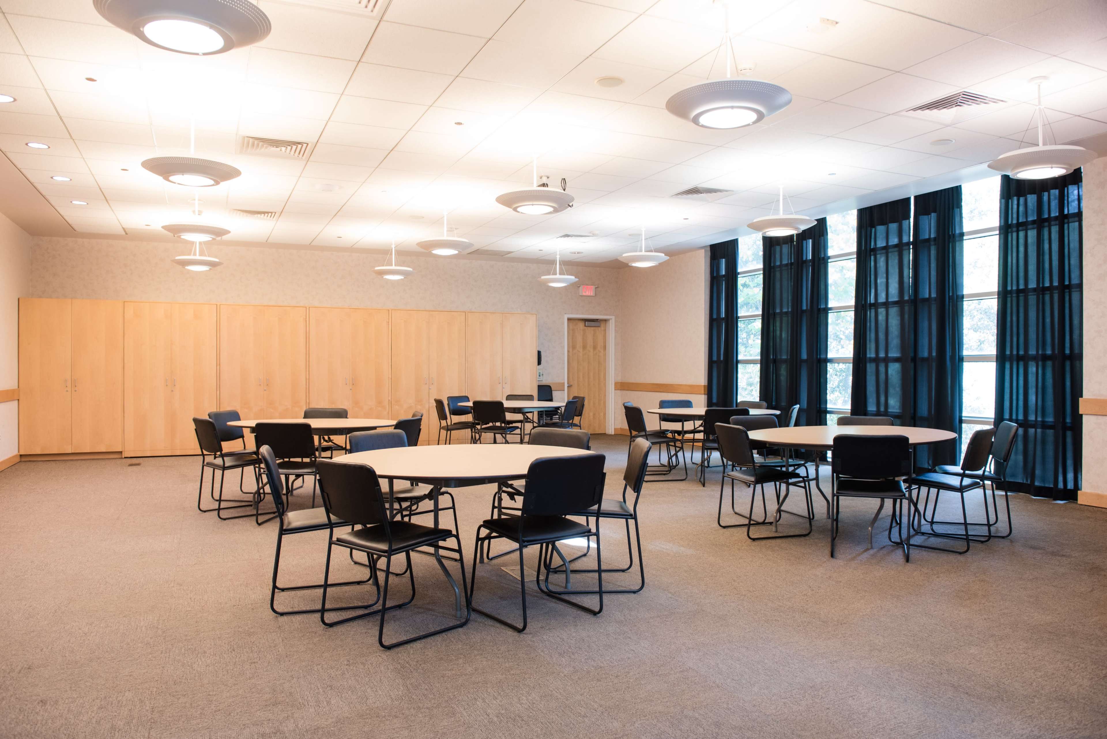 A spacious meeting room featuring several round tables with black chairs arranged around them, illuminated by overhead lights.