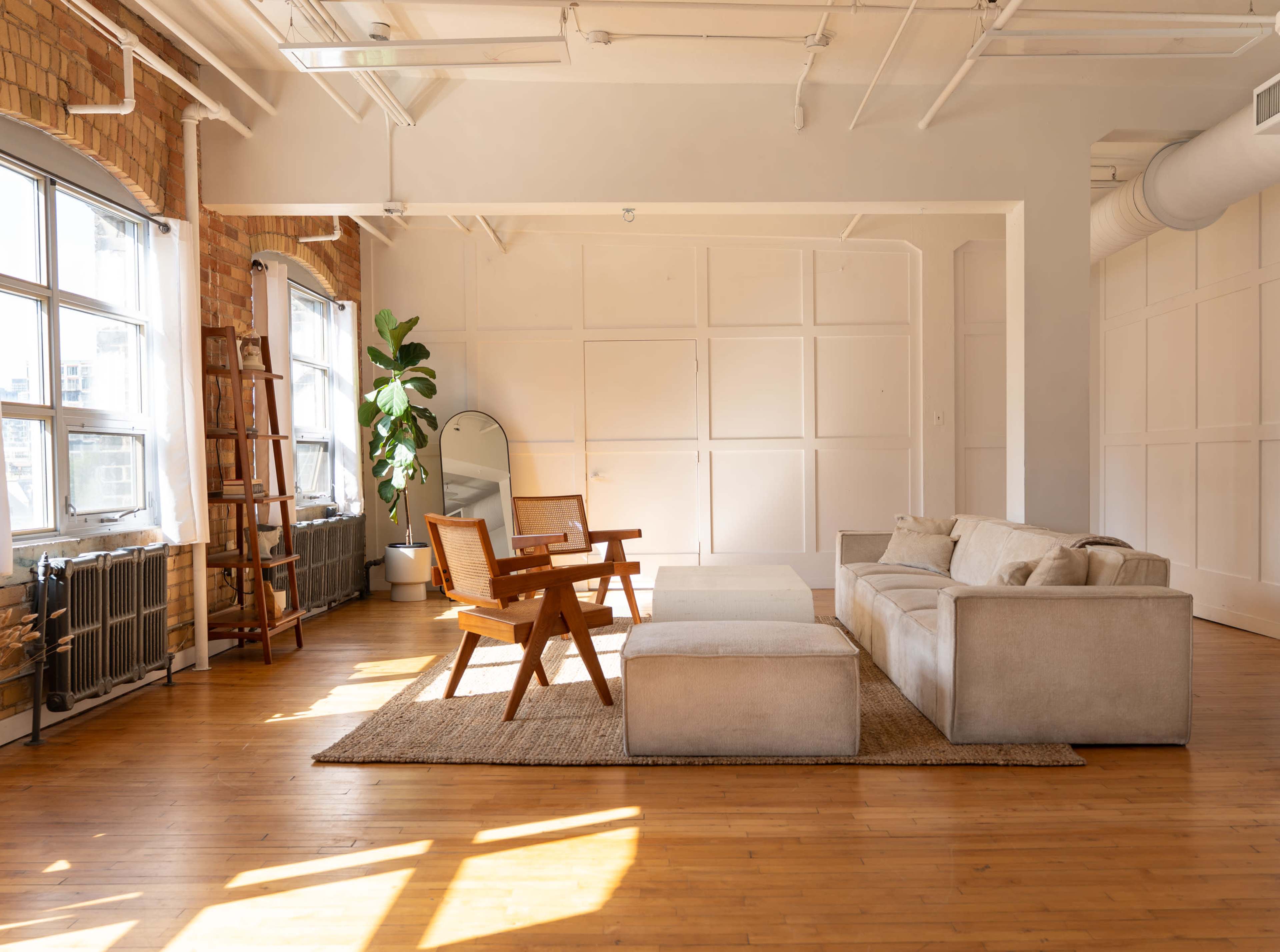 The image shows a bright, spacious living area featuring a light-colored sofa, wooden chairs, a coffee table, and large windows with natural light.