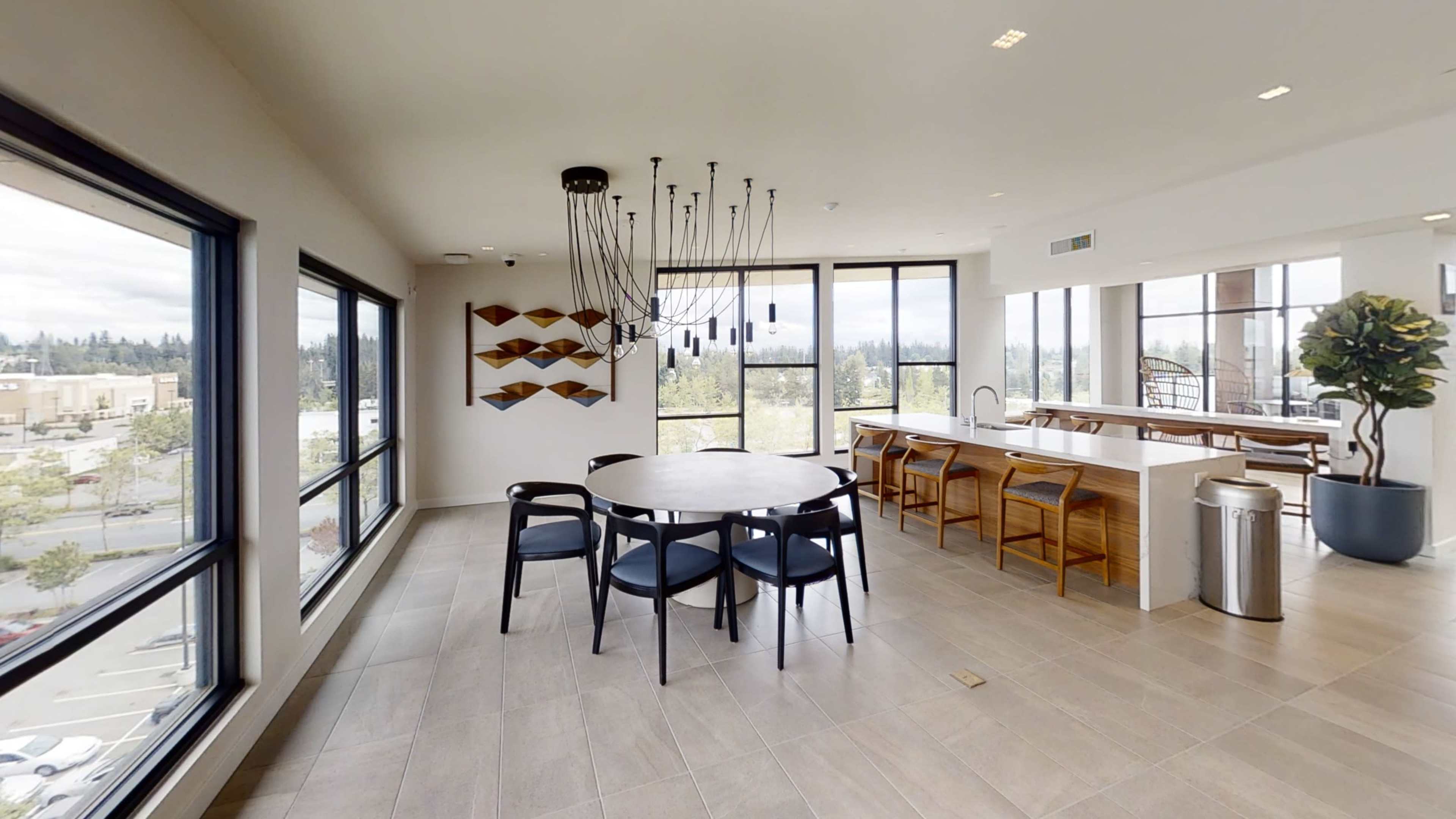 A well-lit communal space featuring a round dining table surrounded by black chairs, large windows, and a kitchen area with wooden elements.