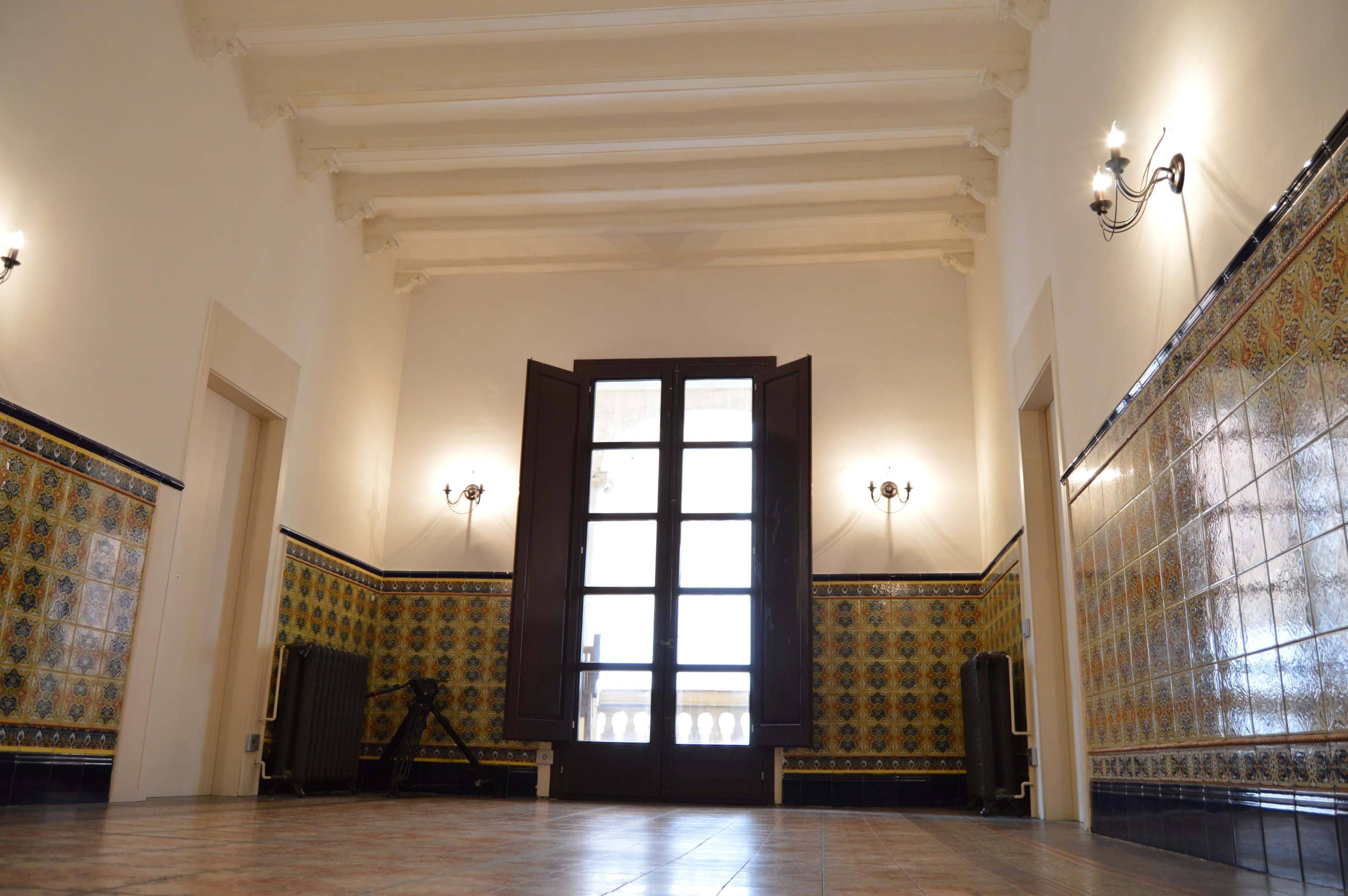 A spacious interior room with patterned tile walls, a set of open wooden doors leading to a balcony, and exposed ceiling beams.