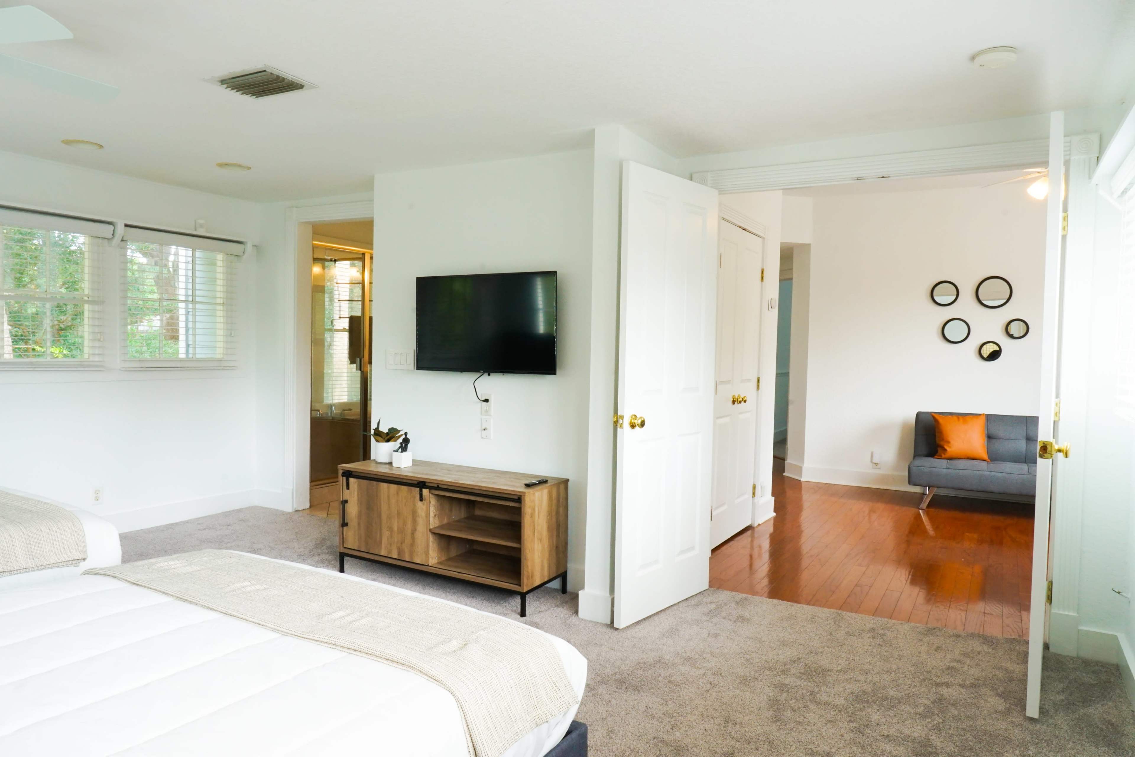 A bedroom with two beds, a television mounted on the wall, a wooden media console, and an open door leading to a living area with a couch and wall art.